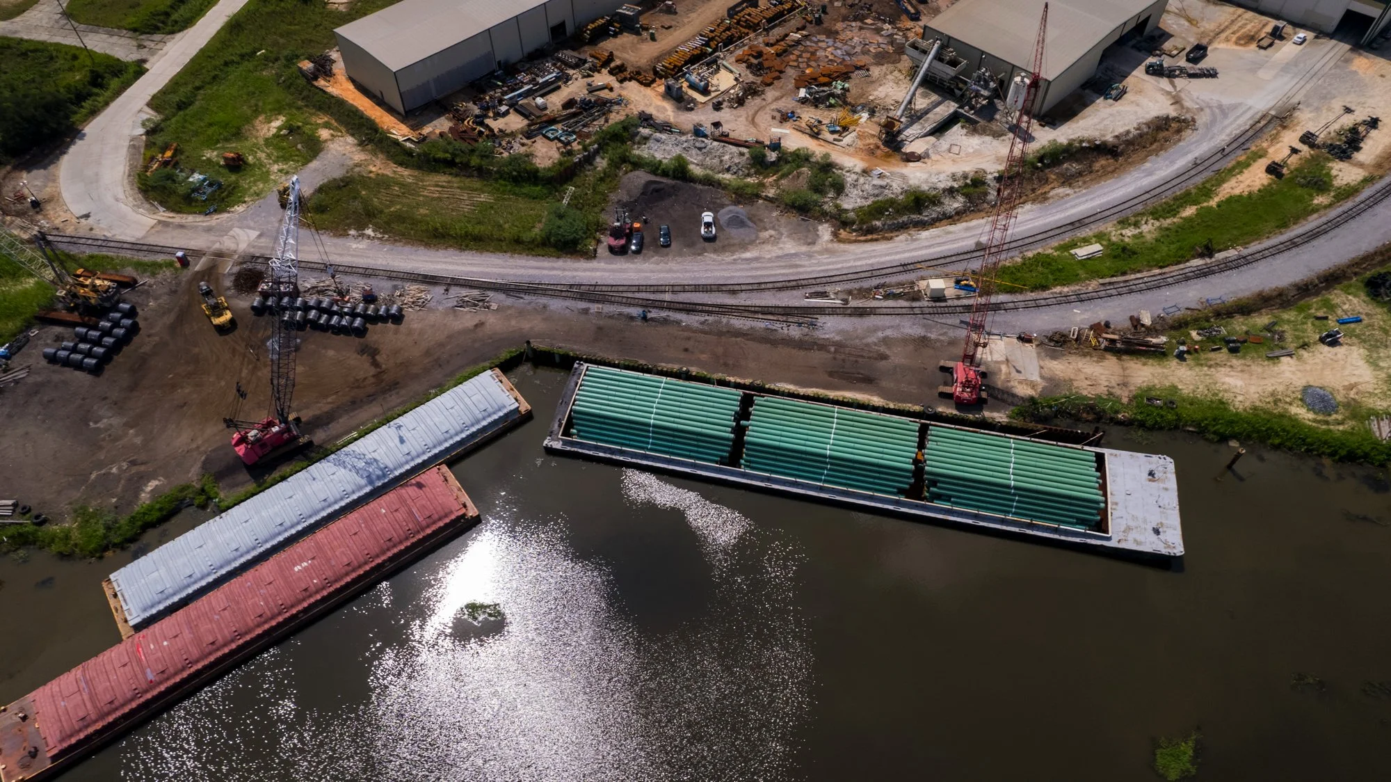 Aerial view of a construction site near water with barges, cranes, construction materials, and parked vehicles.