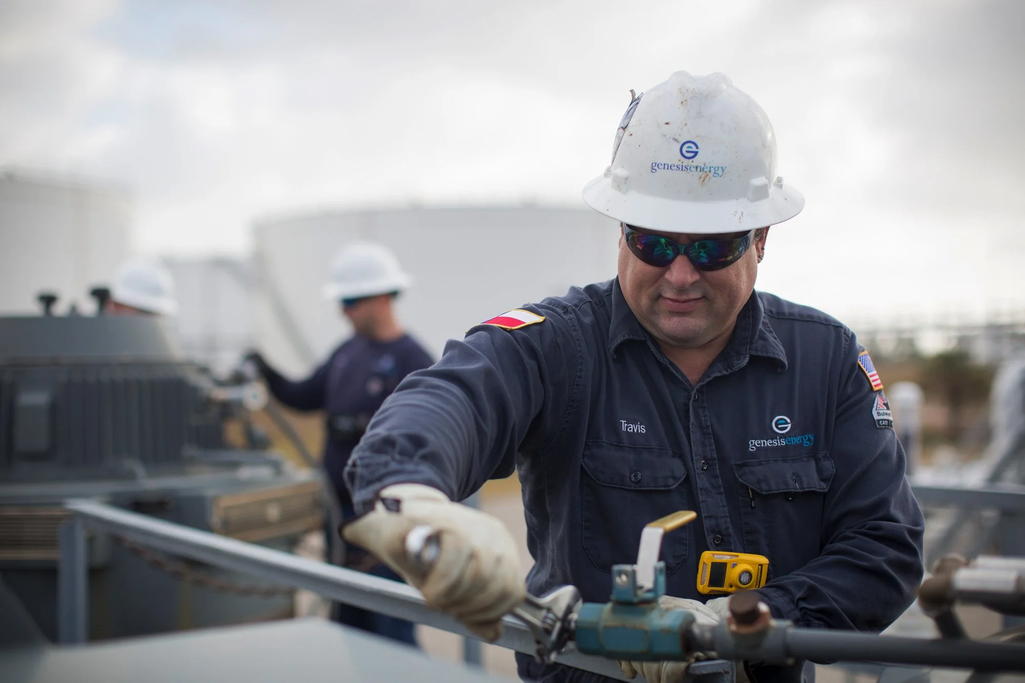 A worker wearing a white Genesis Energy hard hat, sunglasses, and a navy blue uniform with the name 'Travis' on it, working on industrial equipment outdoors.