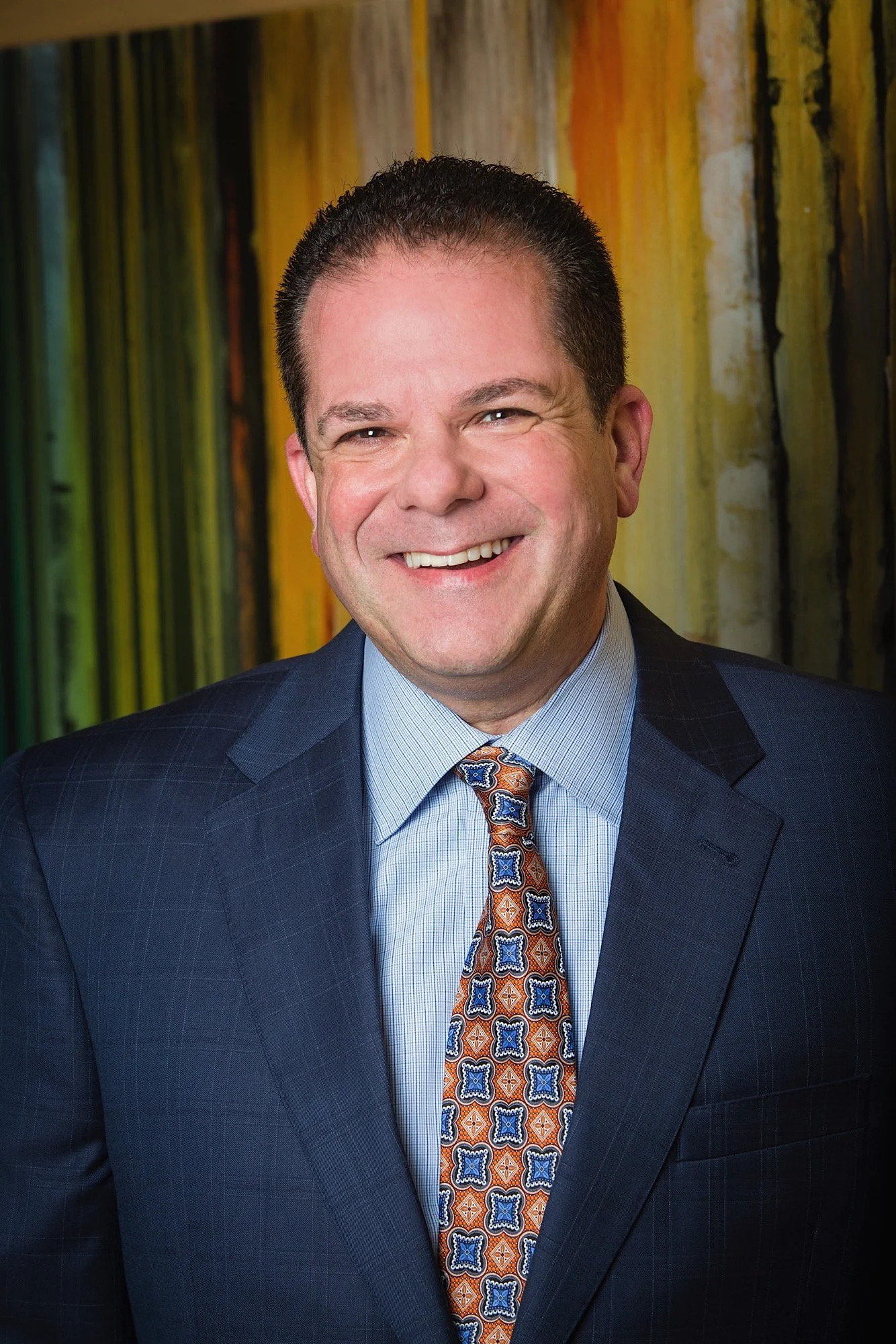A smiling man in a business suit with a patterned tie, standing in front of an abstract colorful background.