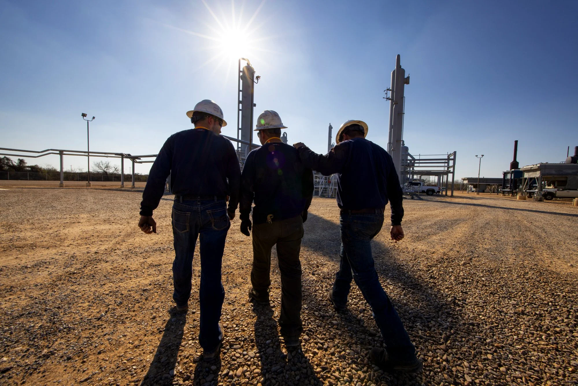 Three workers in hard hats walk together near industrial equipment on a gravel lot under a bright sun.