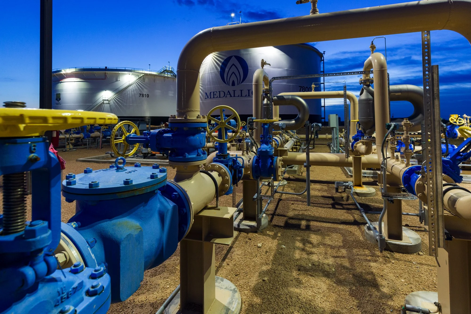 Oil or gas storage tanks with piping and valves at a facility during dusk or dawn with a blue sky.