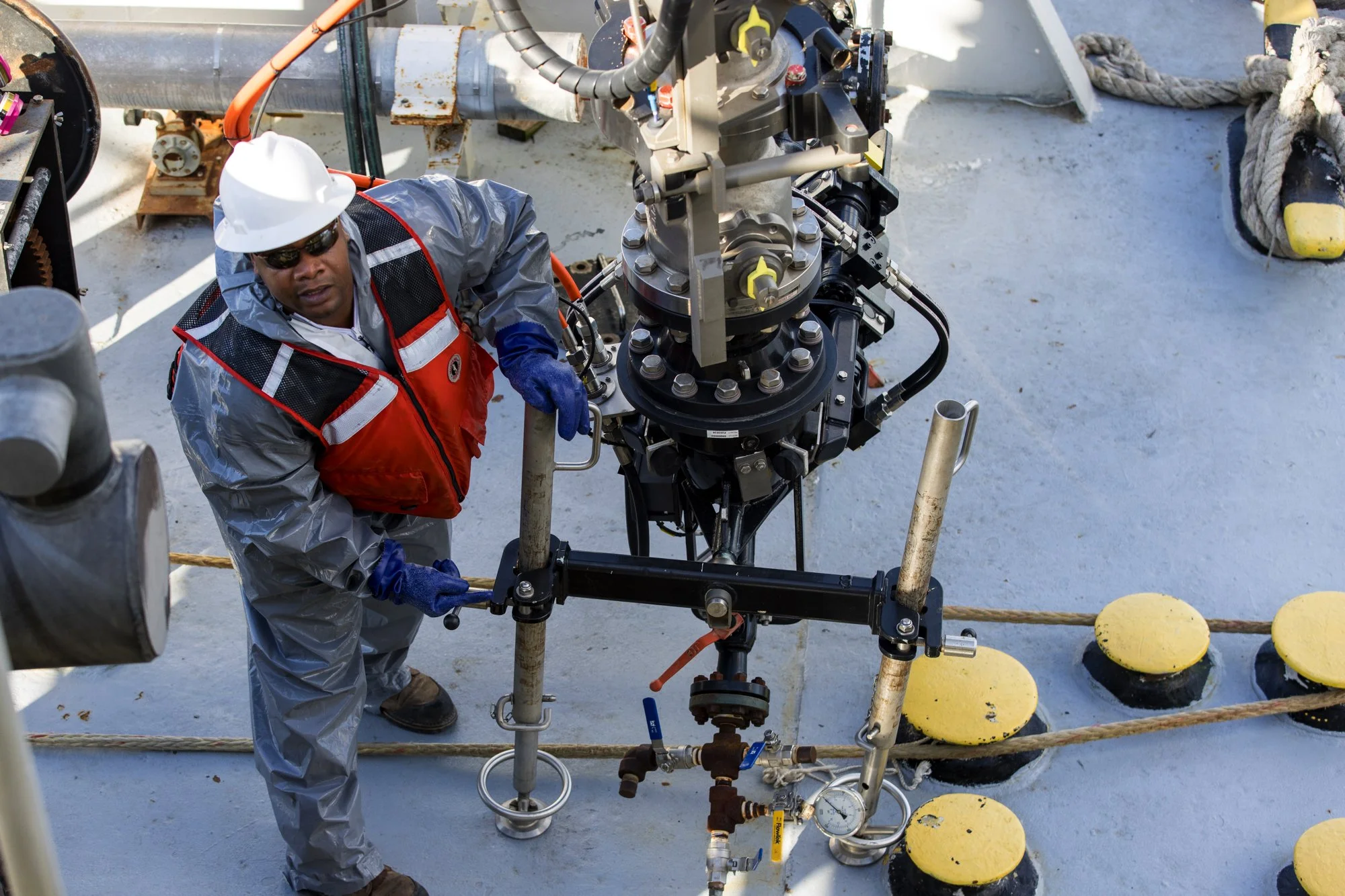 A worker in safety gear, including a helmet, gloves, and a reflective vest, is operating machinery on a ship's deck, which has a blue and gray surface and is surrounded by yellow safety bumpers and thick ropes.