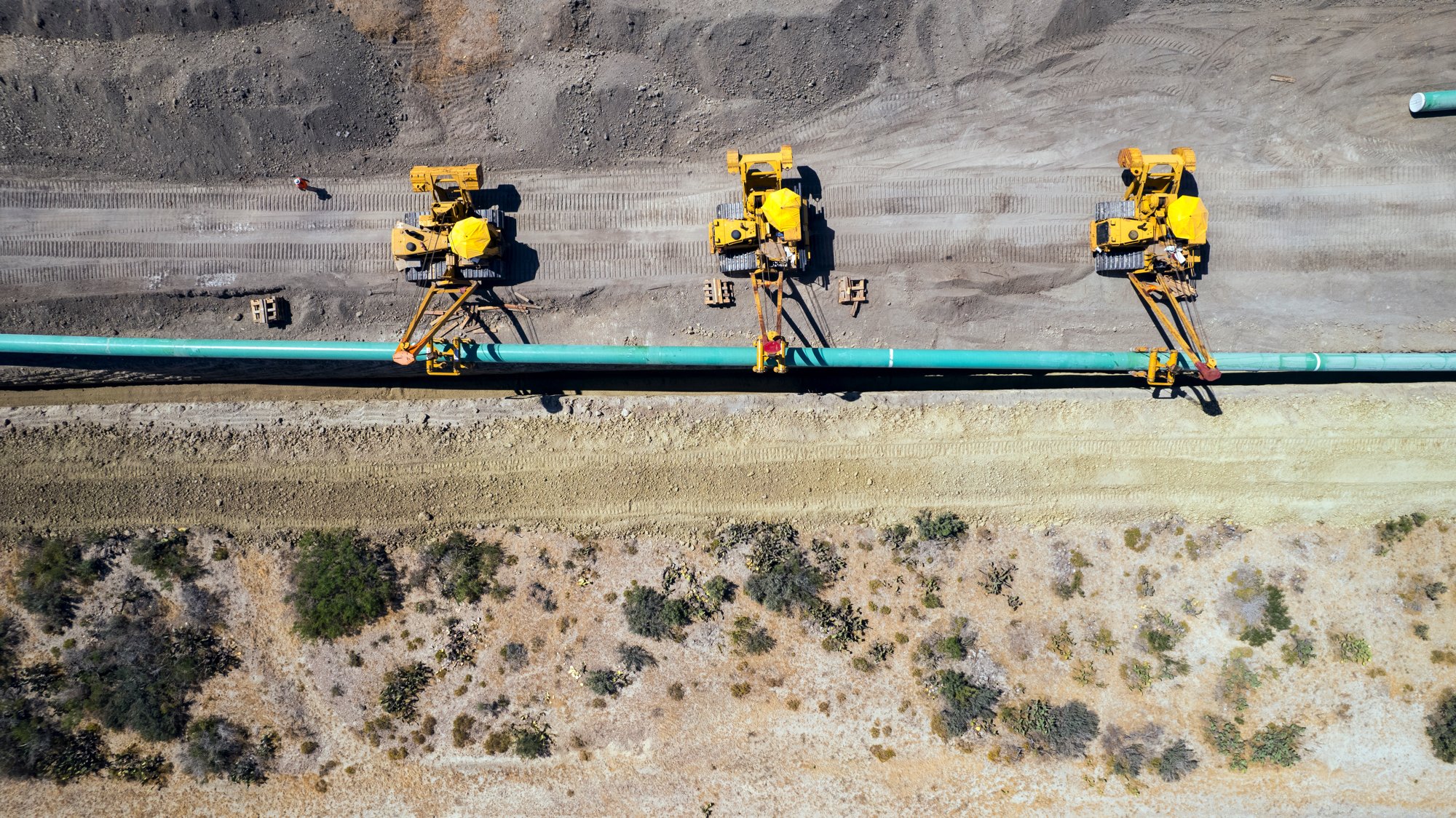 Three large yellow construction machines laying pipeline along a dirt road in a desert-like area with sparse vegetation.