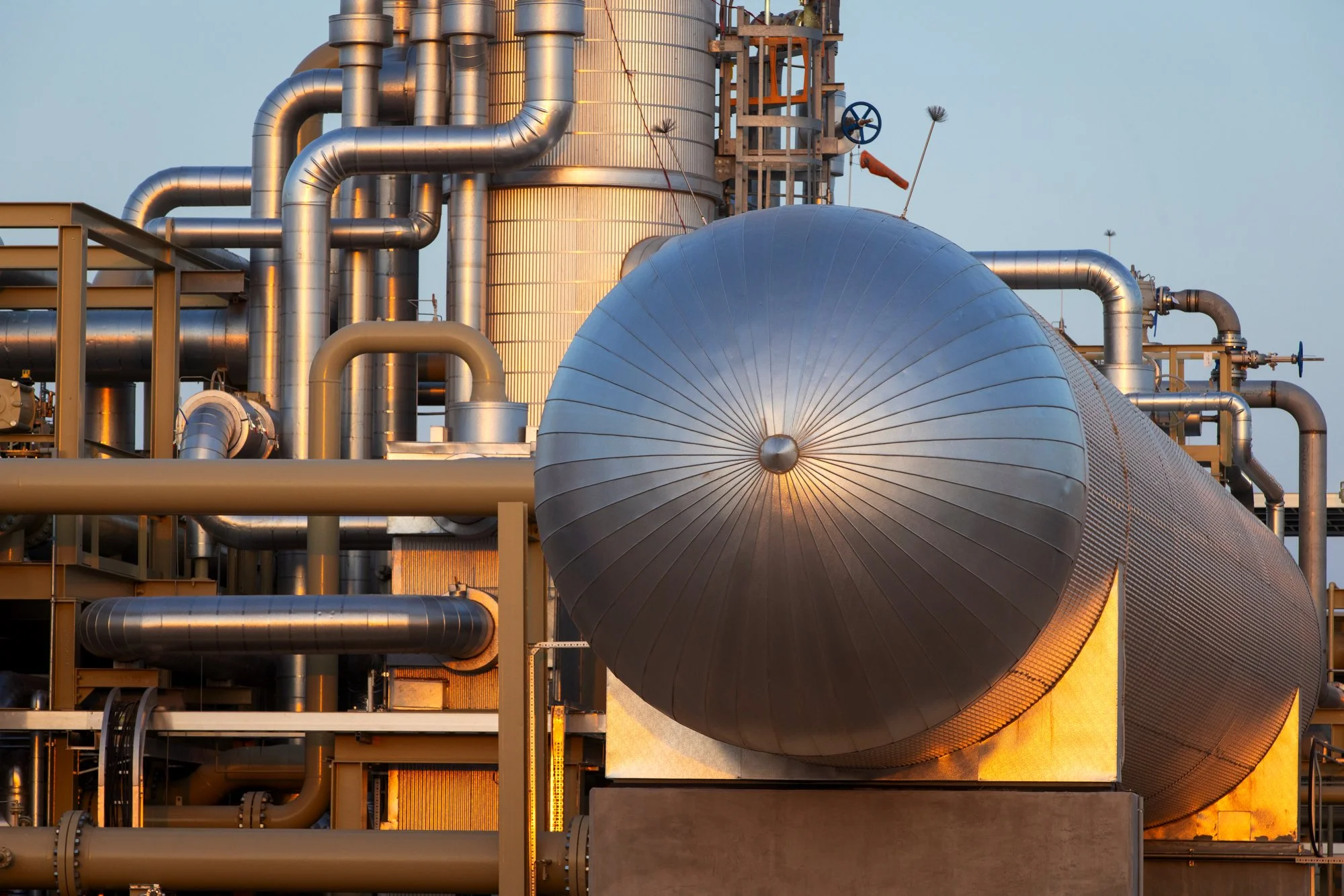 A large industrial facility with numerous silver-colored pipes and a large cylindrical storage tank in the foreground.