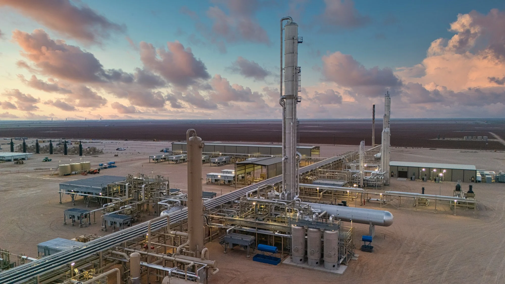 An oil or gas processing facility in a desert landscape with industrial pipes, storage tanks, and structures, under a cloudy sky at sunset.