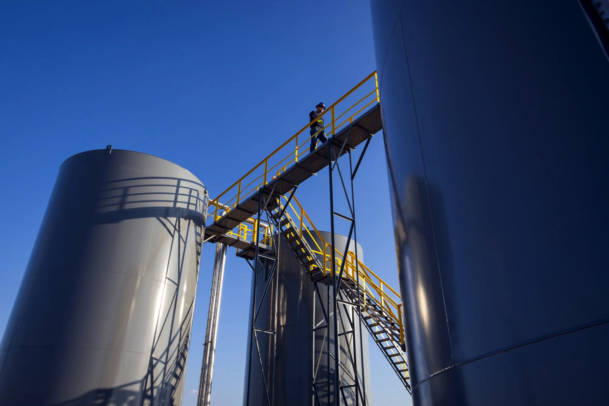 A person in safety gear standing on a metal staircase with yellow railings, adjacent to large silver industrial storage tanks, against a clear blue sky.