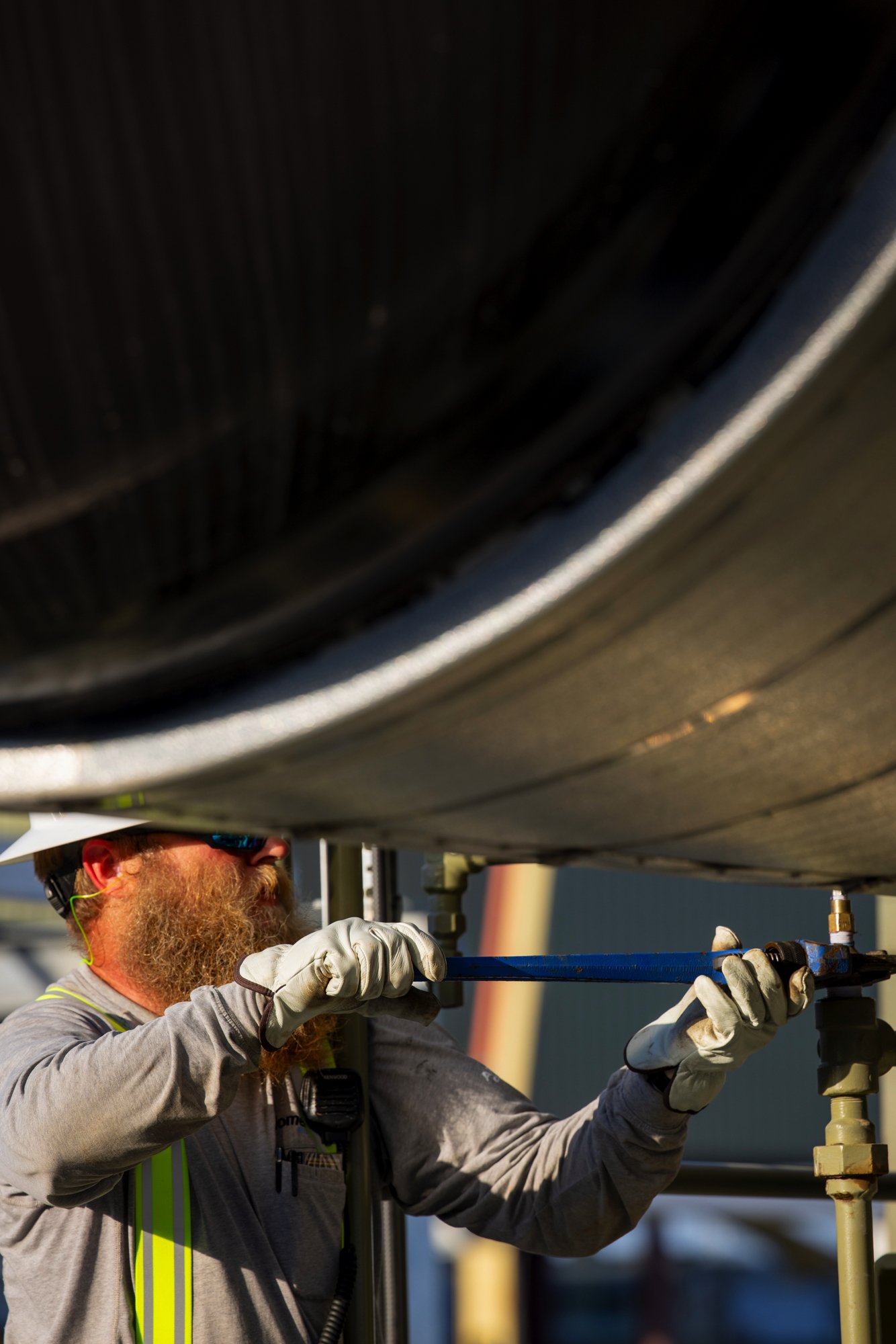 Worker with beard and safety glasses inspecting a pipe outdoors, using a wrench, wearing gloves and a grey shirt.