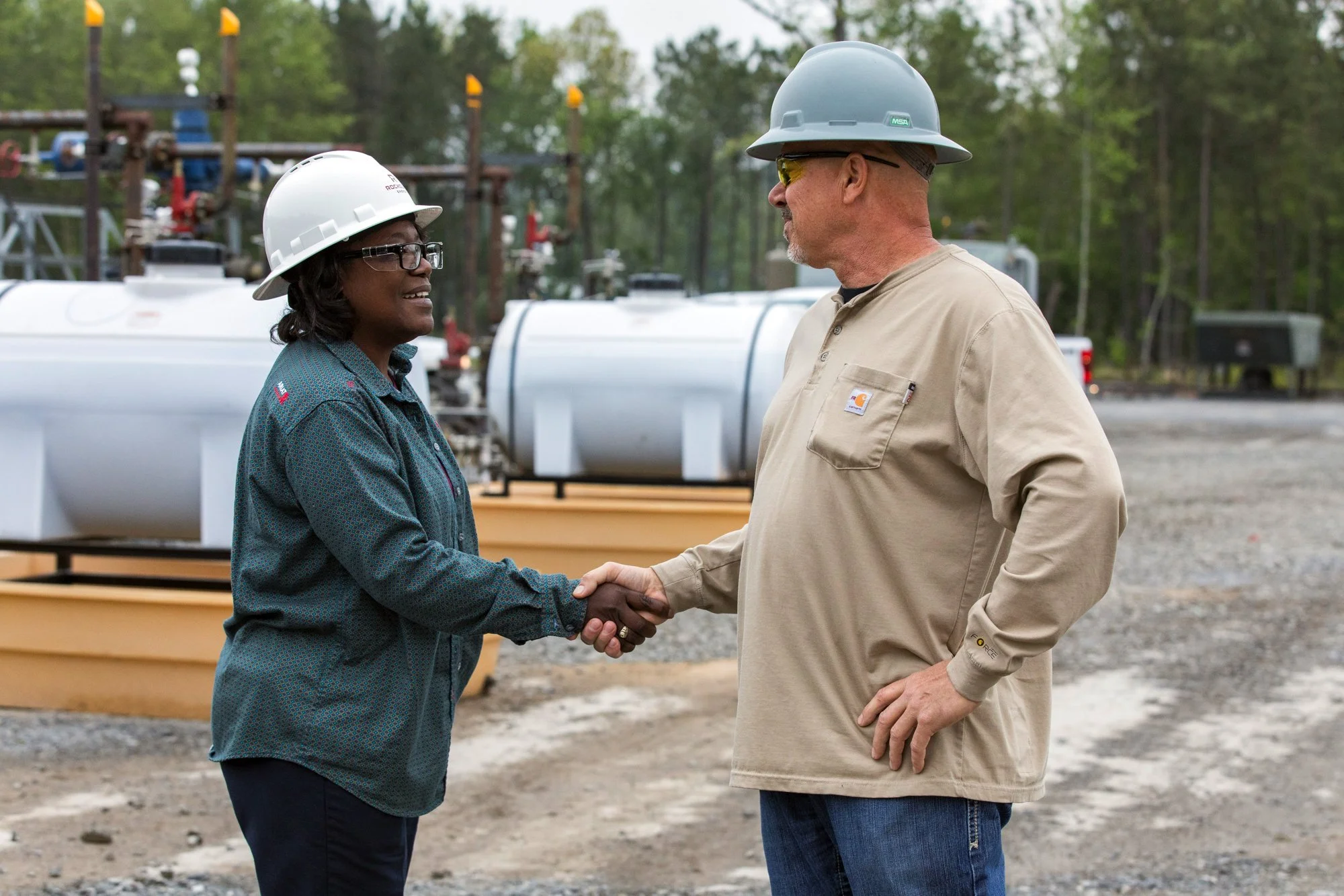 A man and woman wearing safety helmets shaking hands at an outdoor industrial site with pipelines and equipment in the background.