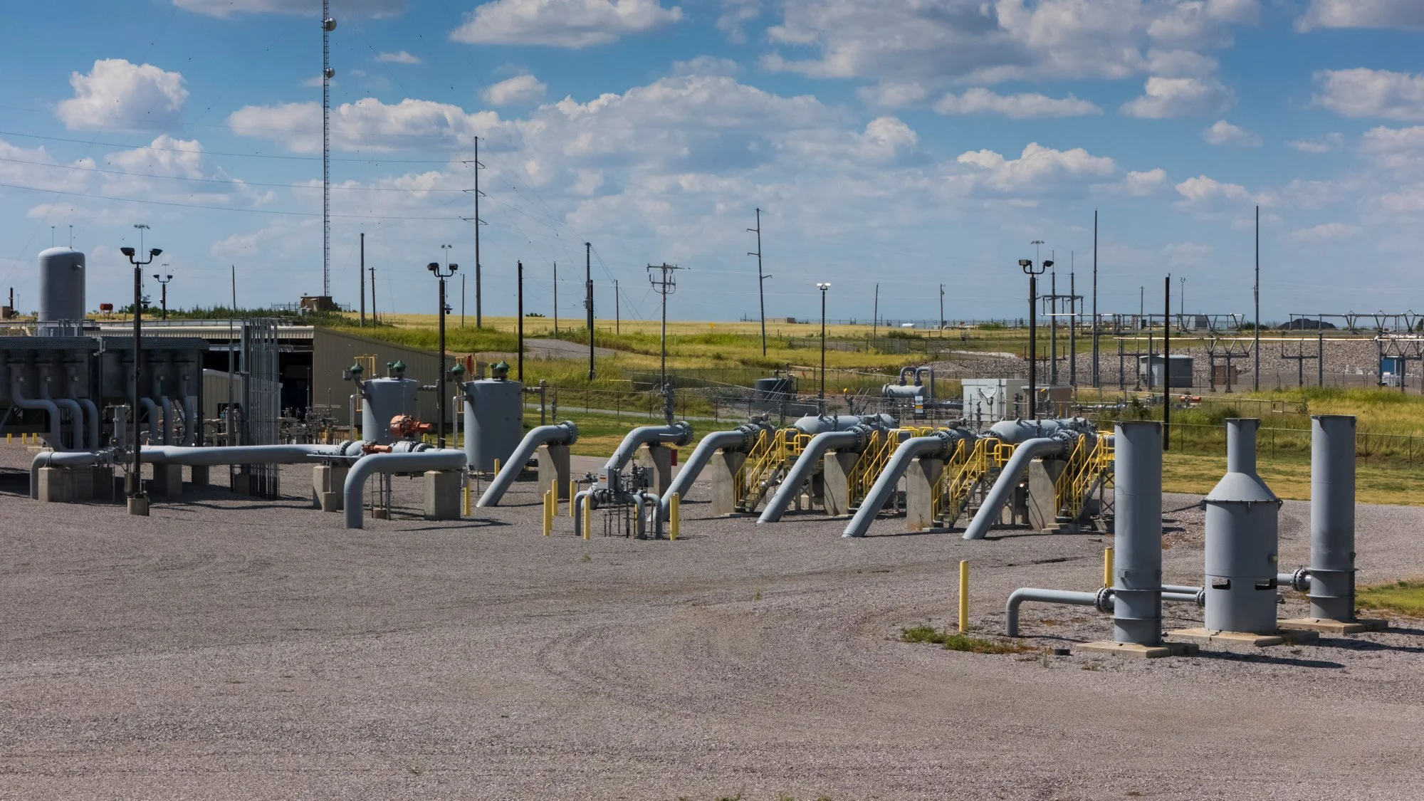 A gas or oil processing facility with pipelines, valves, and control stations outdoors under a blue sky with clouds, surrounded by a grassy landscape and power lines in the background.