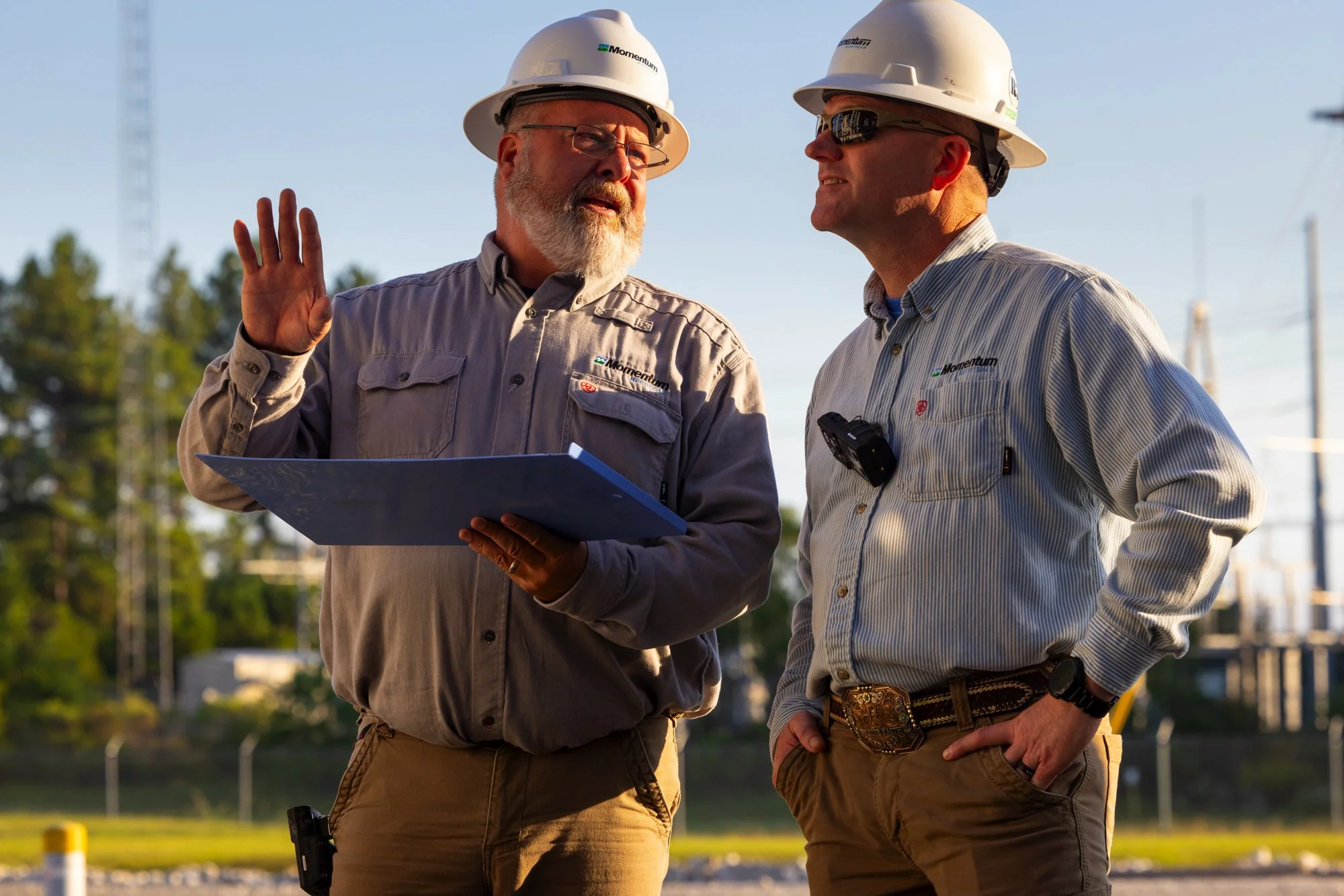 Two male construction workers in hard hats and work shirts having a conversation on a construction site, one holding a blue clipboard.