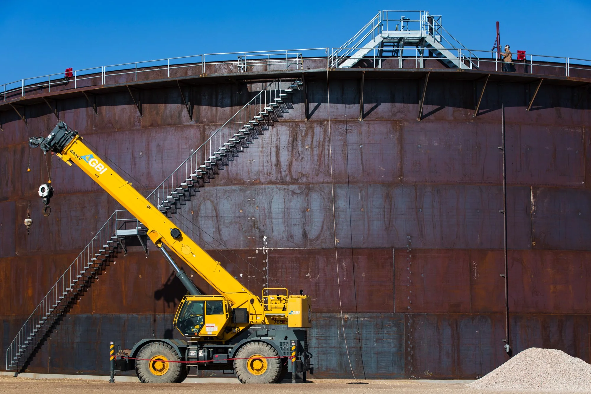 A large yellow crane with the brand GBI is positioned in front of a giant rusted industrial storage tank. The crane's boom is raised and extended, with a hook hanging down. There are external staircase structures on the tank leading to a platform at 