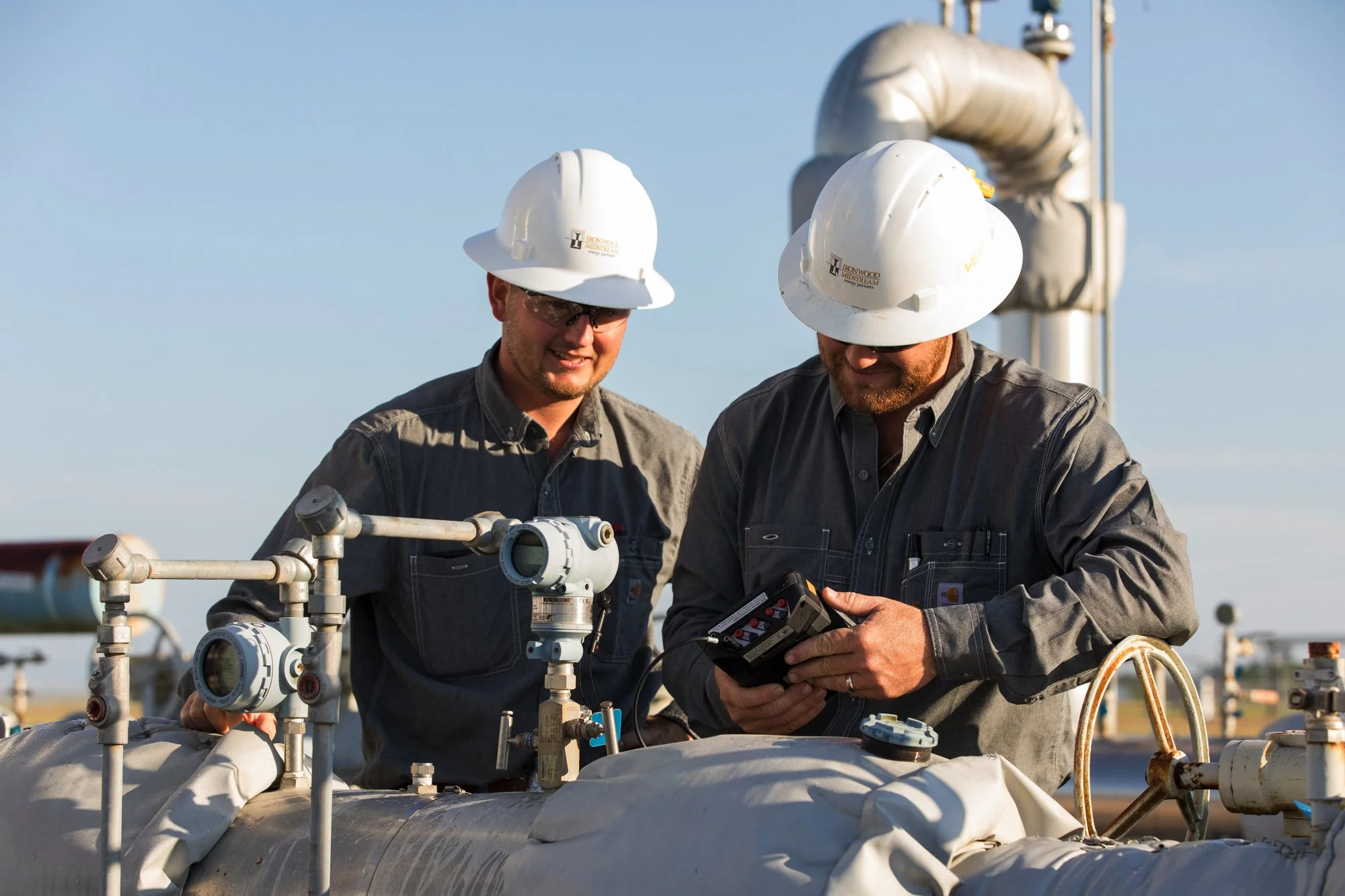 Two male workers wearing white safety helmets and gray work overalls inspecting equipment on a pipeline outdoors.