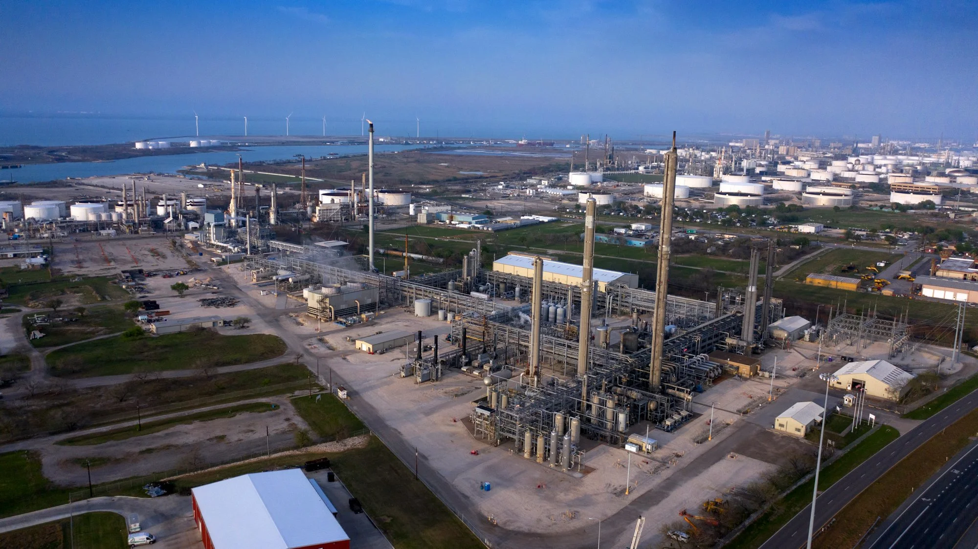 Aerial view of an industrial oil refinery with multiple distillation towers, storage tanks, and pipelines, adjacent to a water body and wind turbines in the background.