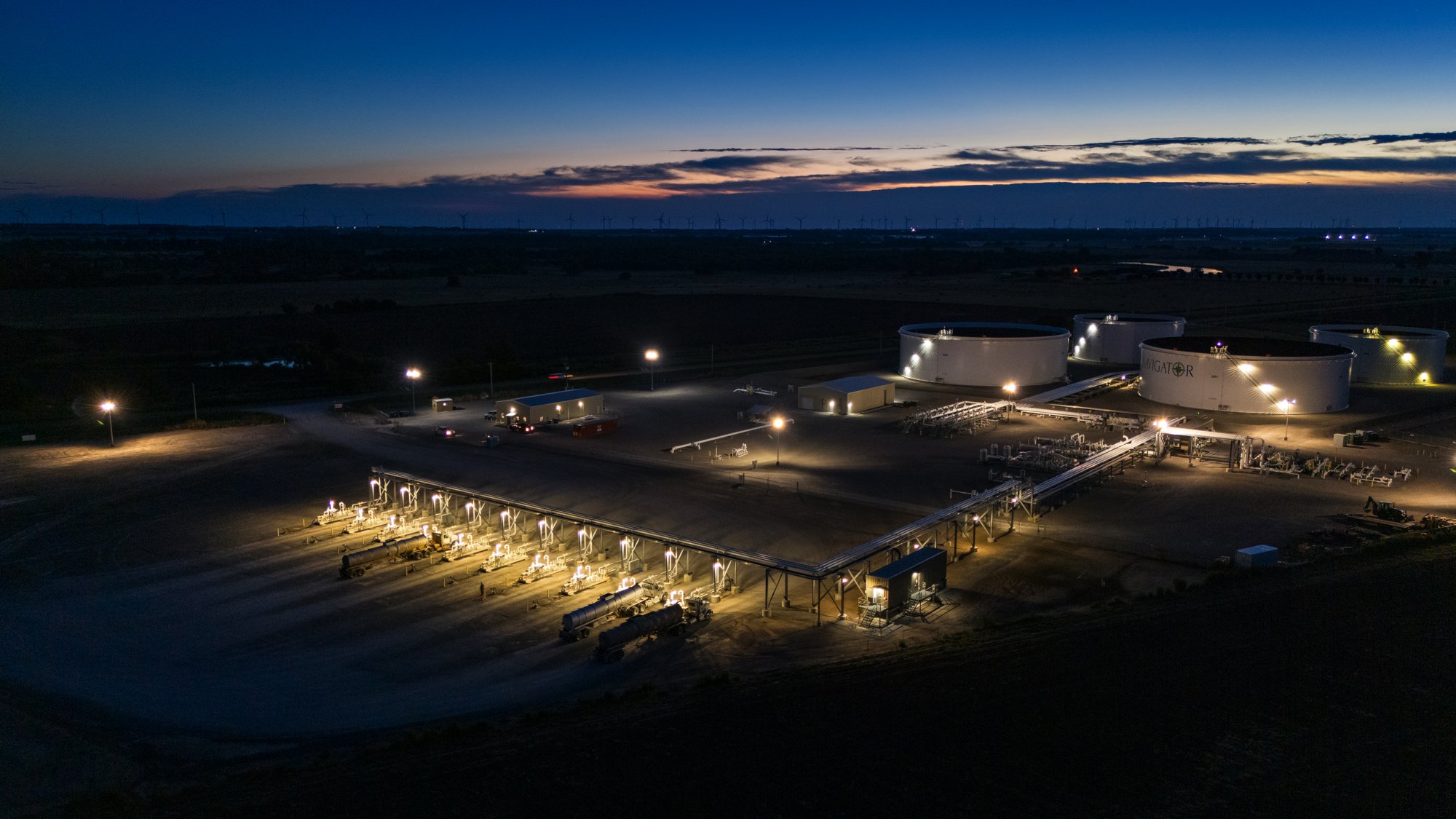 An aerial night view of an industrial fuel storage facility with large tanks, pipelines, and illuminated pump stations, set against a dark landscape with wind turbines on the horizon.