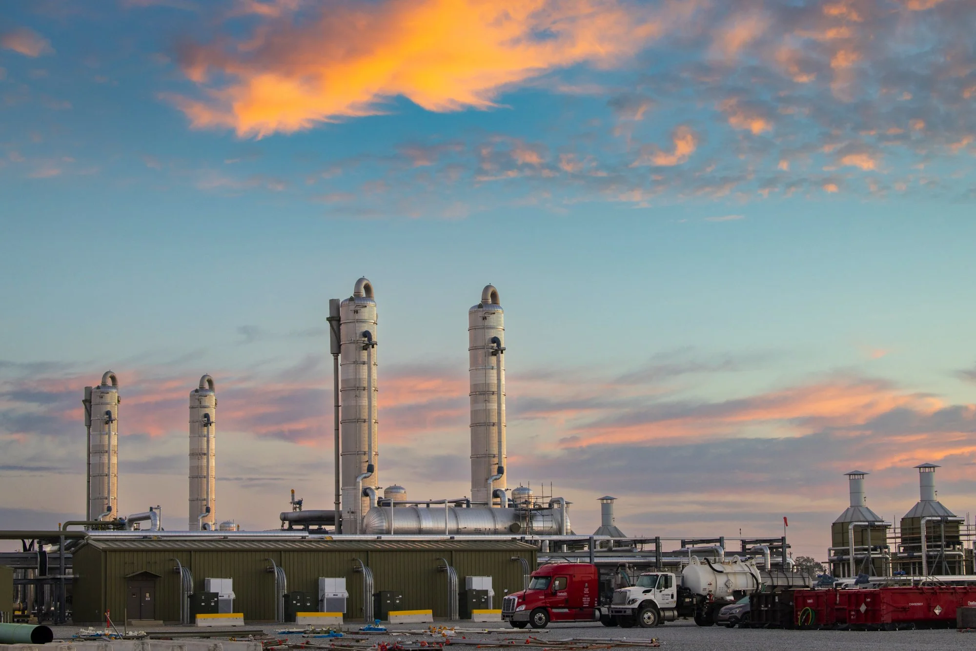 An industrial facility with four tall white smokestacks, a green building with small doors and pipes, and vehicles including a red semi-truck and white tanker truck, set against a sunset sky with pink and blue clouds.
