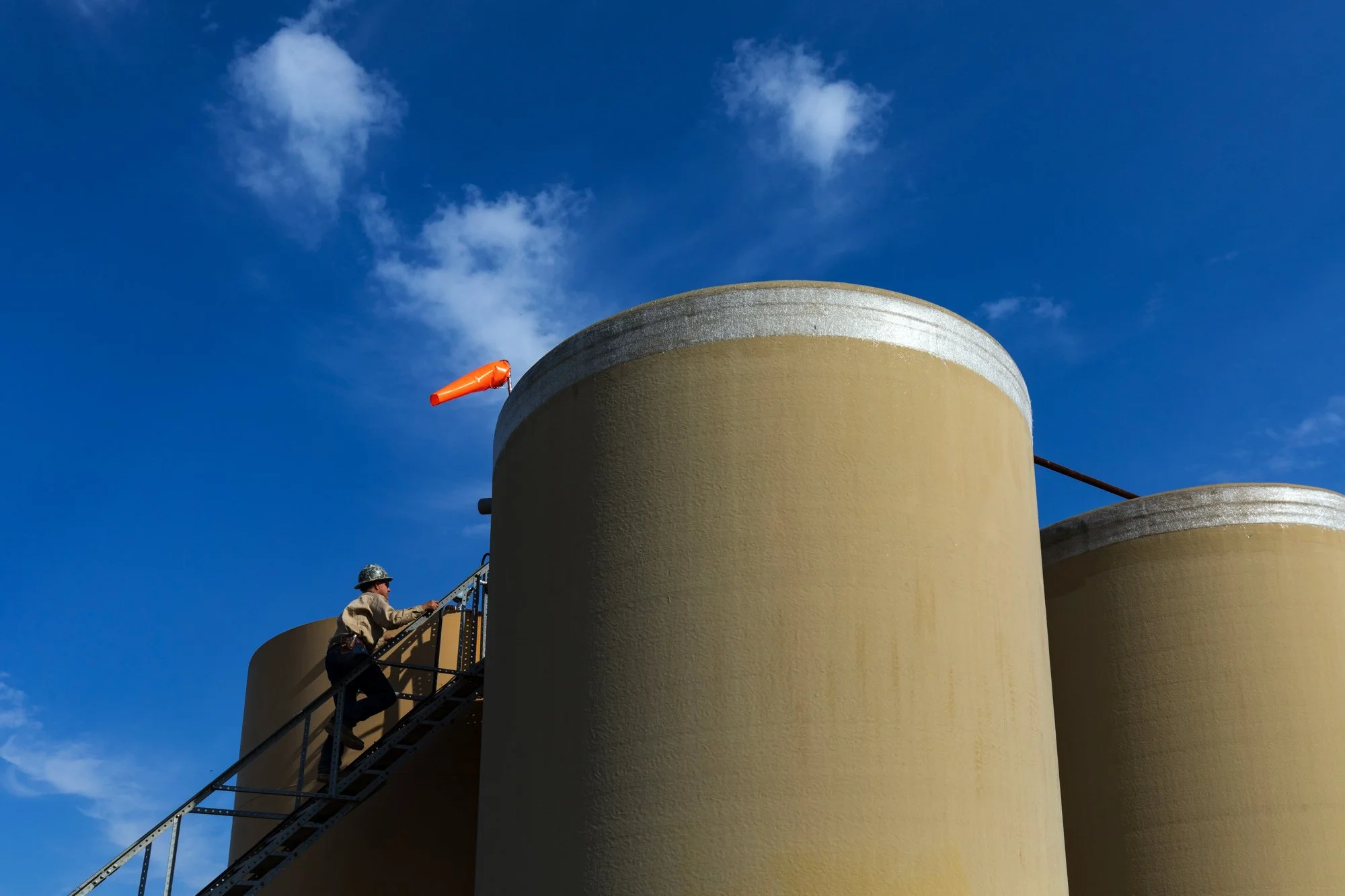 A person in safety gear ascending a metal staircase attached to large beige industrial tanks under a blue sky with some clouds.