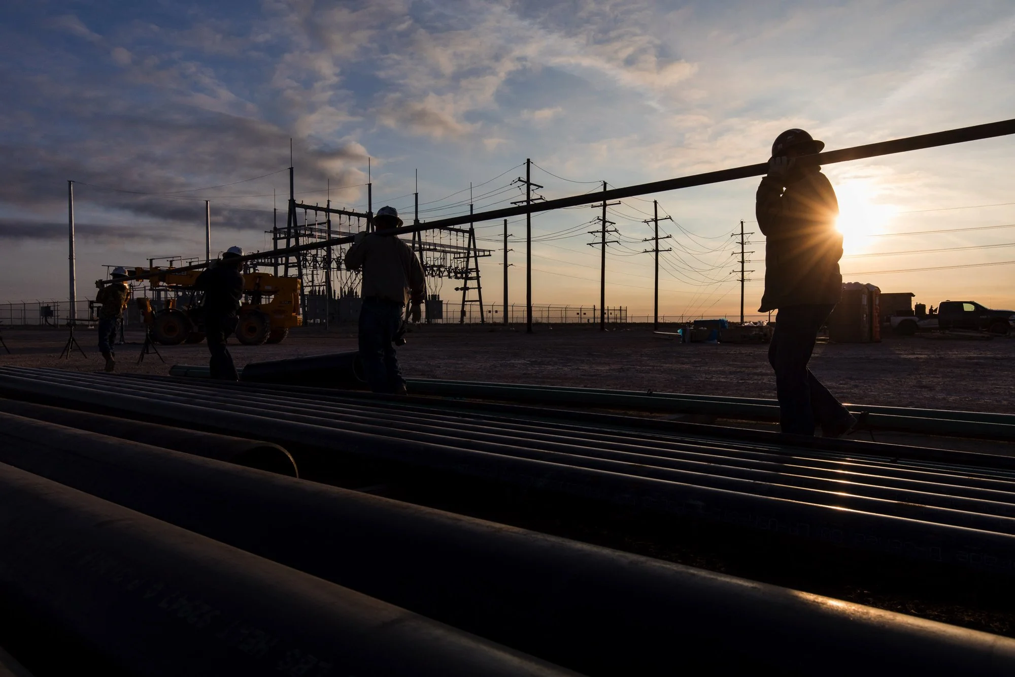 Construction workers working on pipelines outdoors at sunset, with power lines and a utility truck in the background.