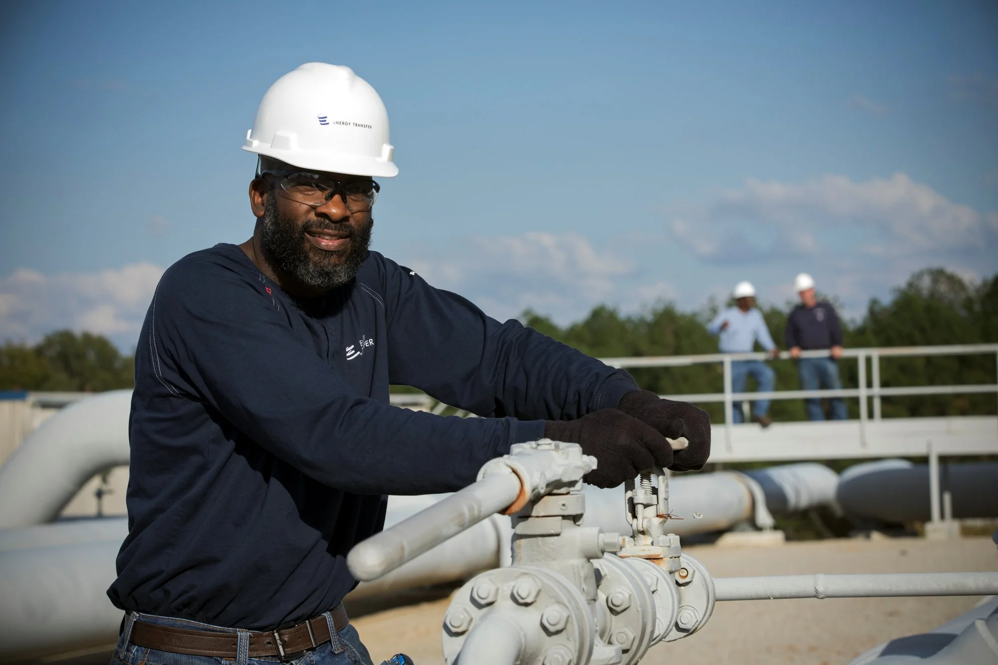 A worker wearing a white safety helmet, glasses, and gloves, operating valves on a pipeline at an industrial site with two other workers in the background.