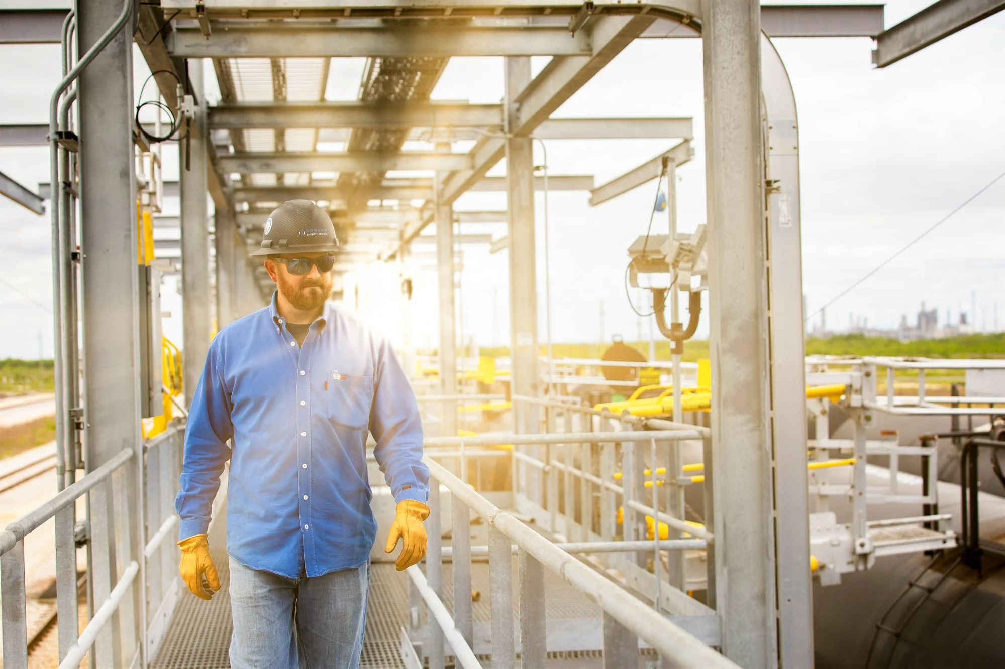 A man in a blue work shirt, yellow gloves, sunglasses, and a hard hat walking on a metal platform at a construction or industrial site with a bright light shining behind him.