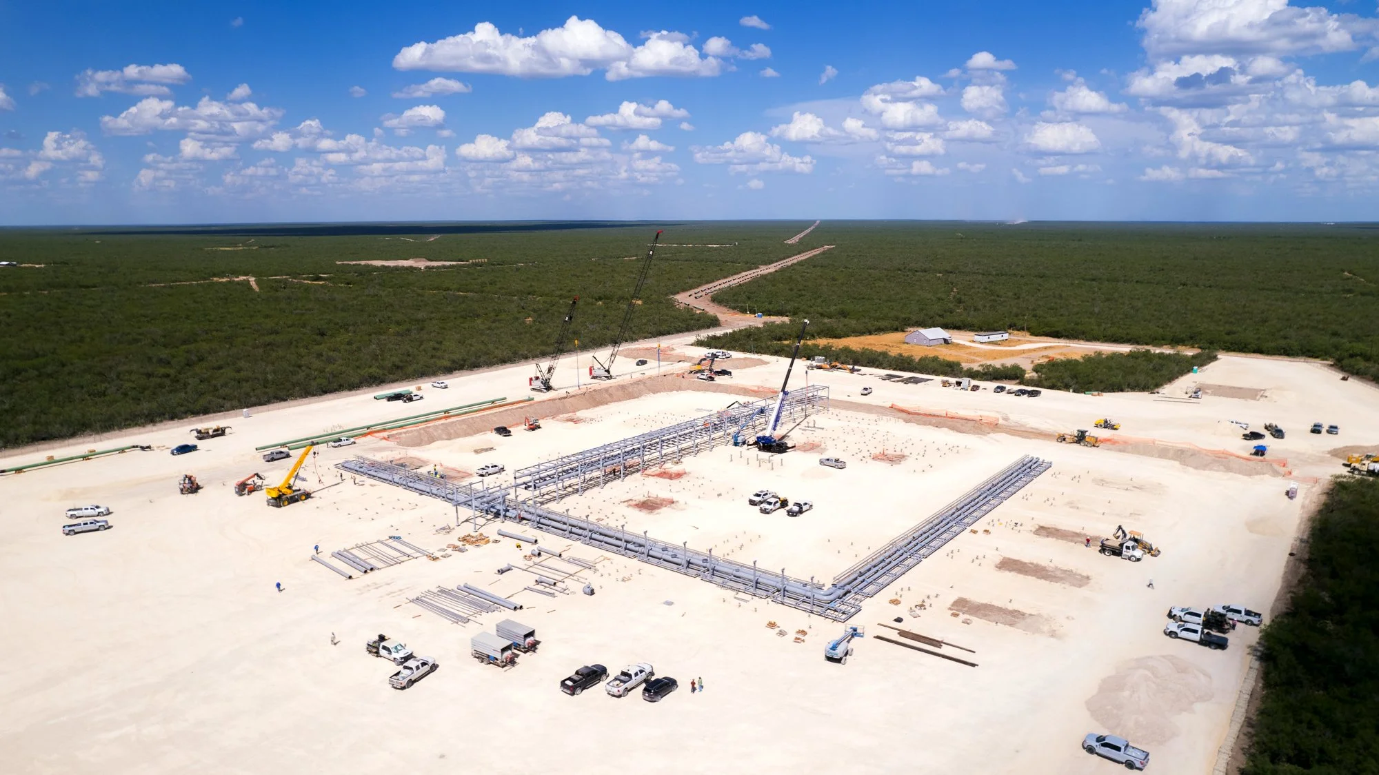Construction site in a large, flat, rural area with several cranes, trucks, and workers building a large framework structure, surrounded by green forest under a partly cloudy sky.