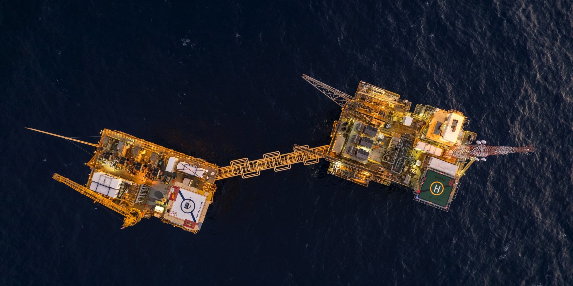 An aerial view of an offshore oil platform with yellow structures, connected by a bridge, surrounded by deep blue ocean.