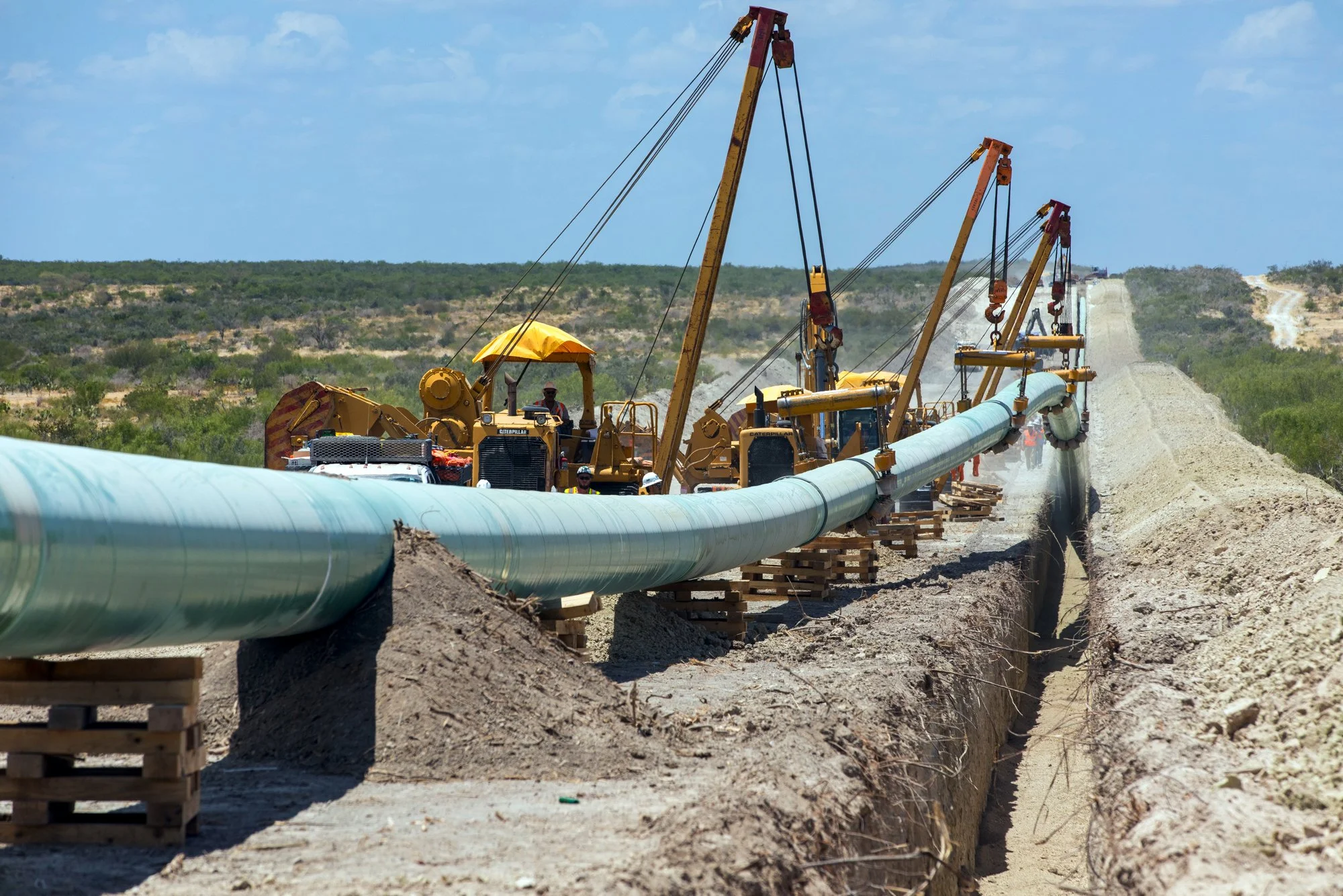 Construction workers using machinery to lay large pipeline across a desert landscape with a dirt road in the distance and clear blue sky.