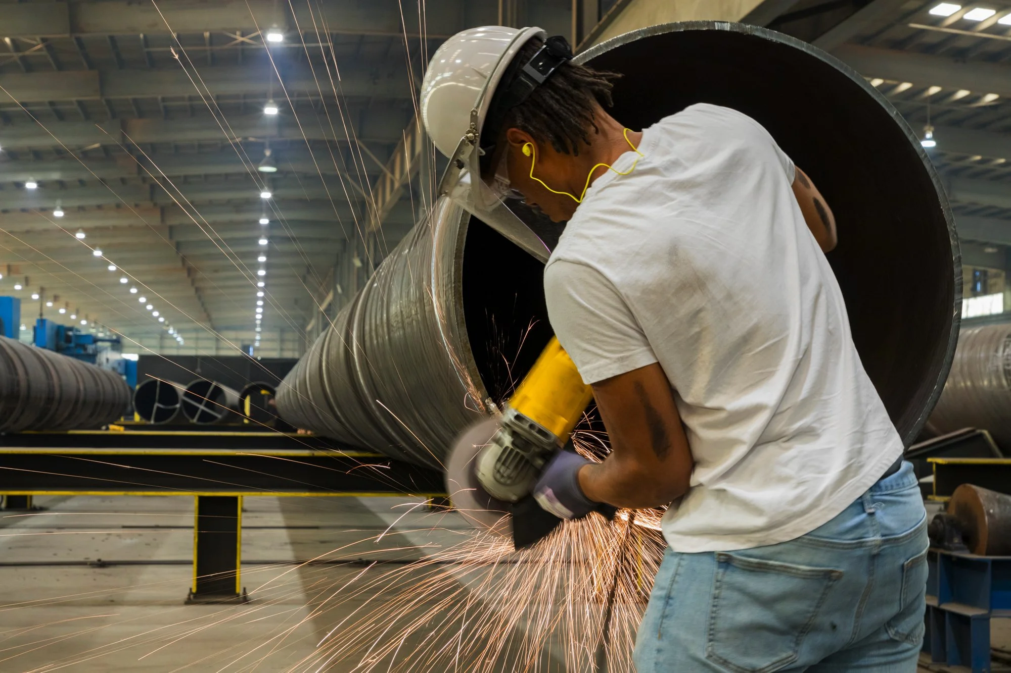 Worker cutting metal pipe with a grinder, sparks flying in a manufacturing or industrial facility.
