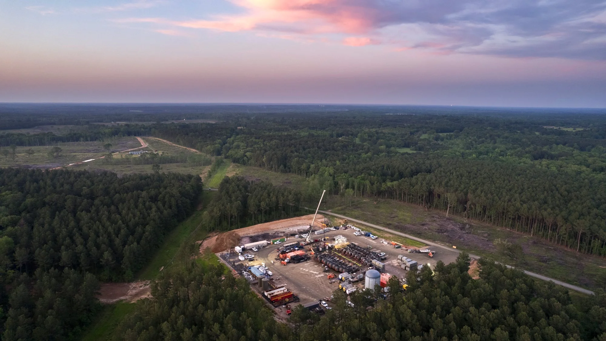 An aerial view of a construction site in a forested area during dusk, with construction equipment, vehicles, and a crane present.