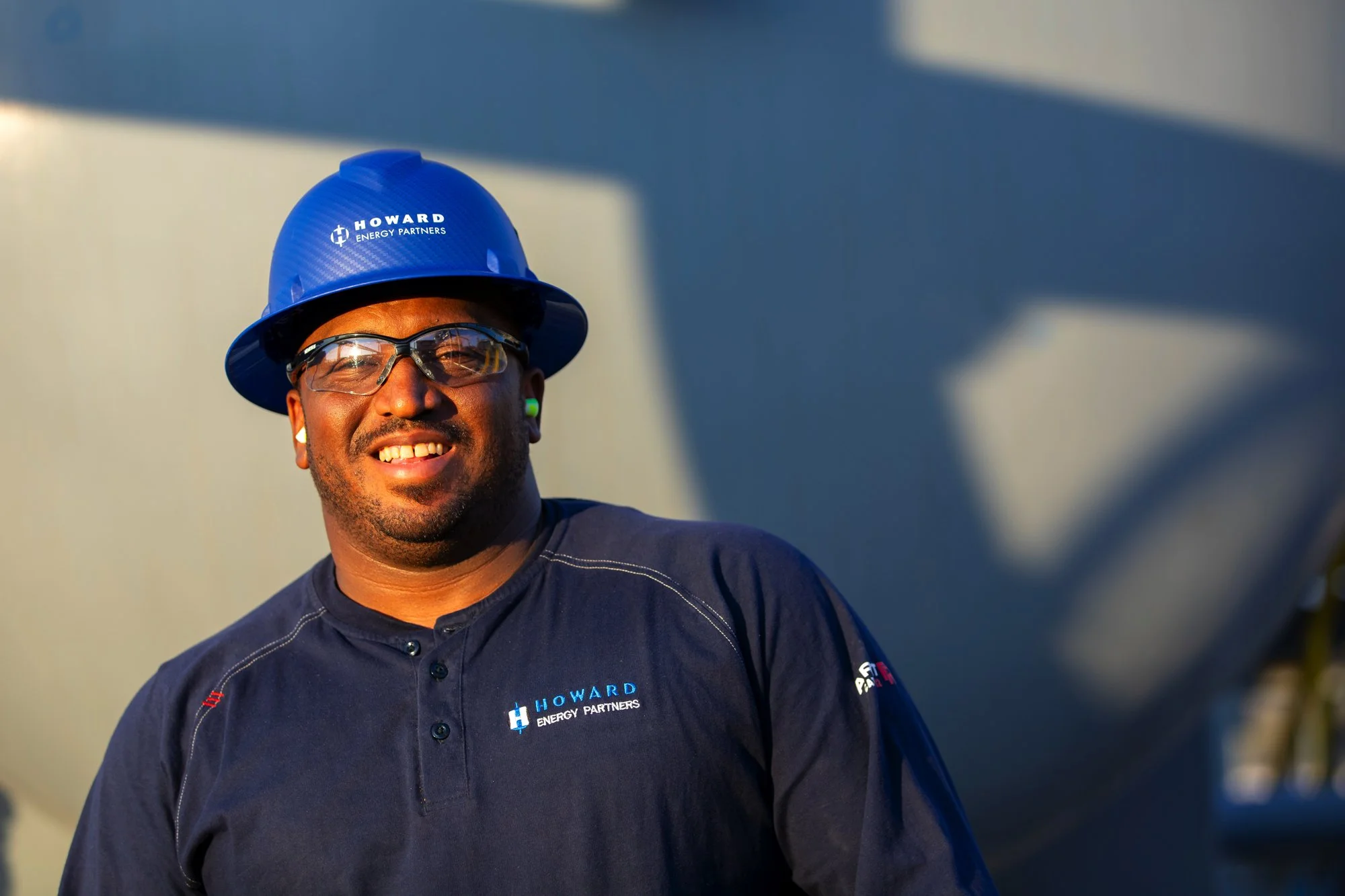 A smiling man with glasses and earrings wearing a blue hard hat and a dark shirt with the logo 'Howard Energy Partners'.