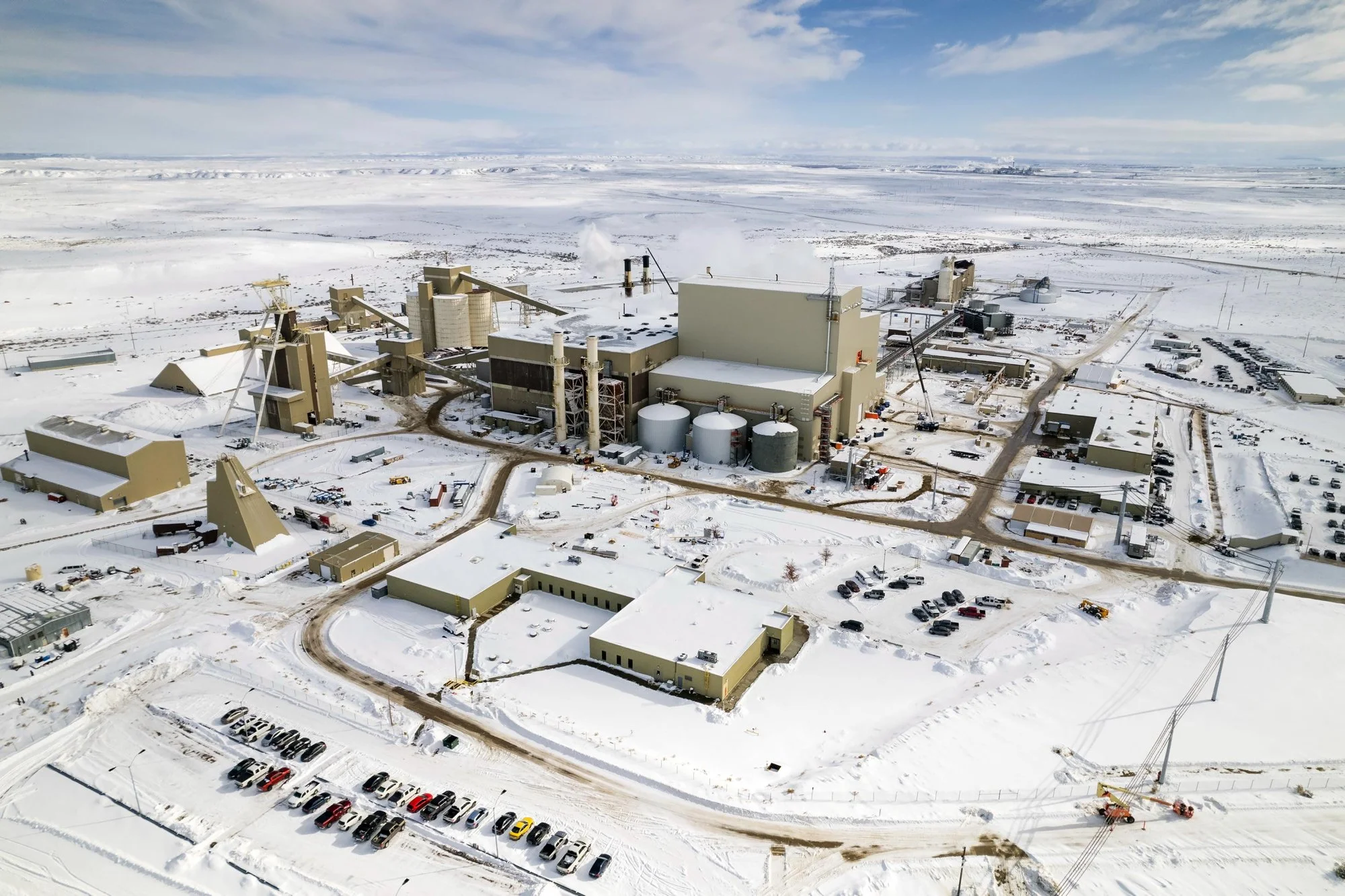 Aerial view of a large industrial complex in a snowy landscape, with multiple buildings, storage tanks, cranes, and parked cars.