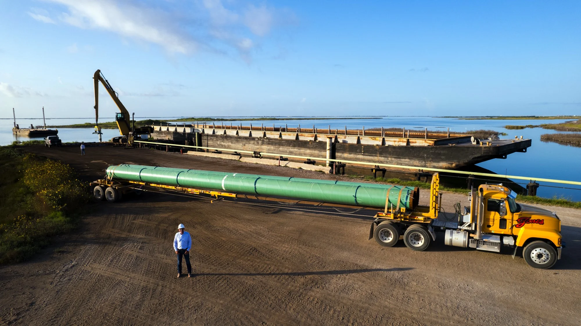 Construction site by a waterway with large equipment, a barge, tall crane, and a yellow truck carrying a large green pipe.