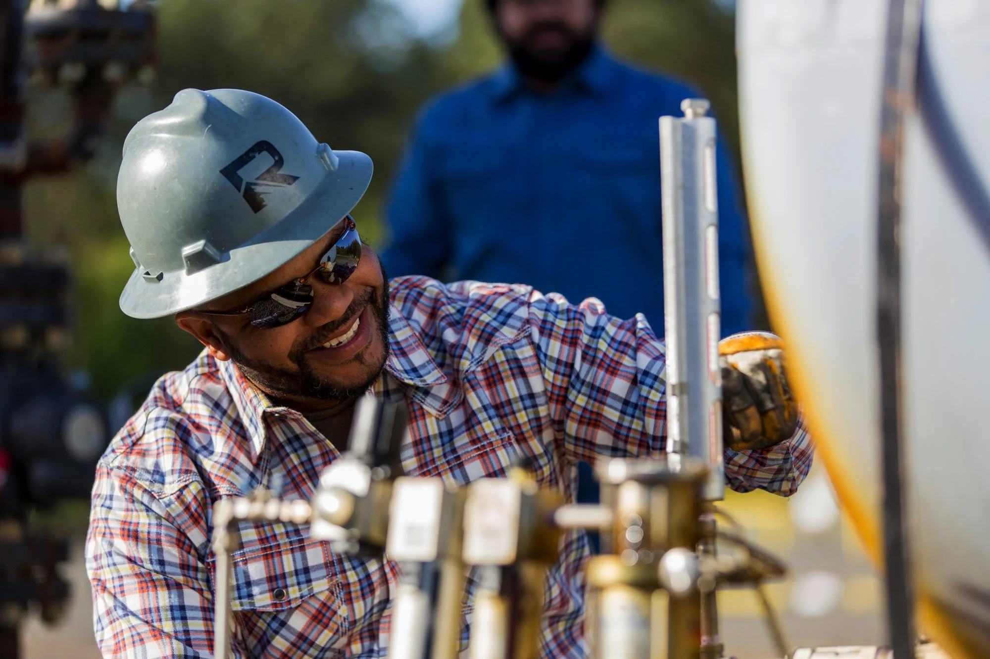 A smiling construction worker wearing a safety helmet, sunglasses, and a plaid shirt, working with metal equipment outdoors, with another worker in the background.