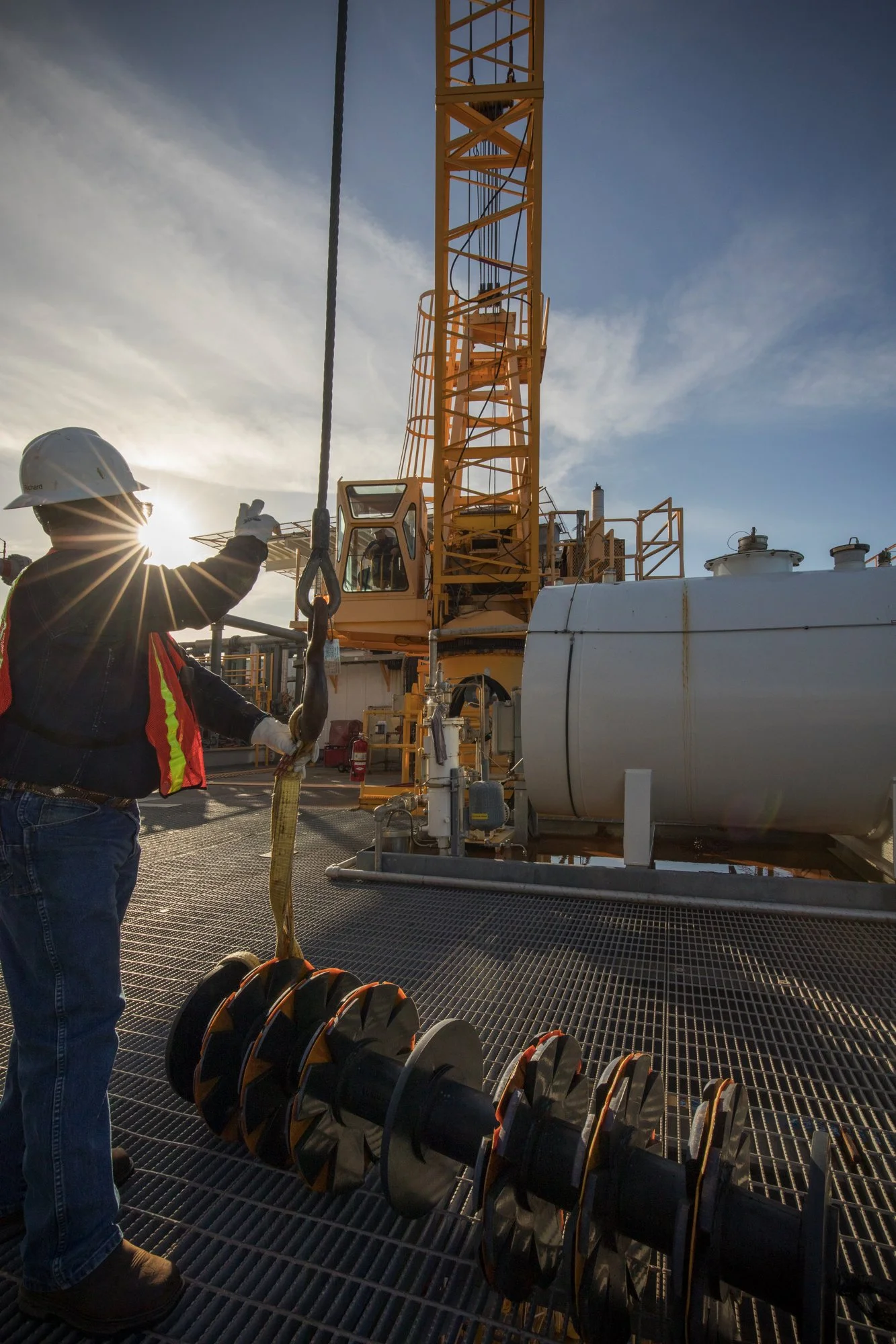 A worker with a hard hat and safety vest on an oil rig, handling equipment attached to a crane with a large white tank in the background.