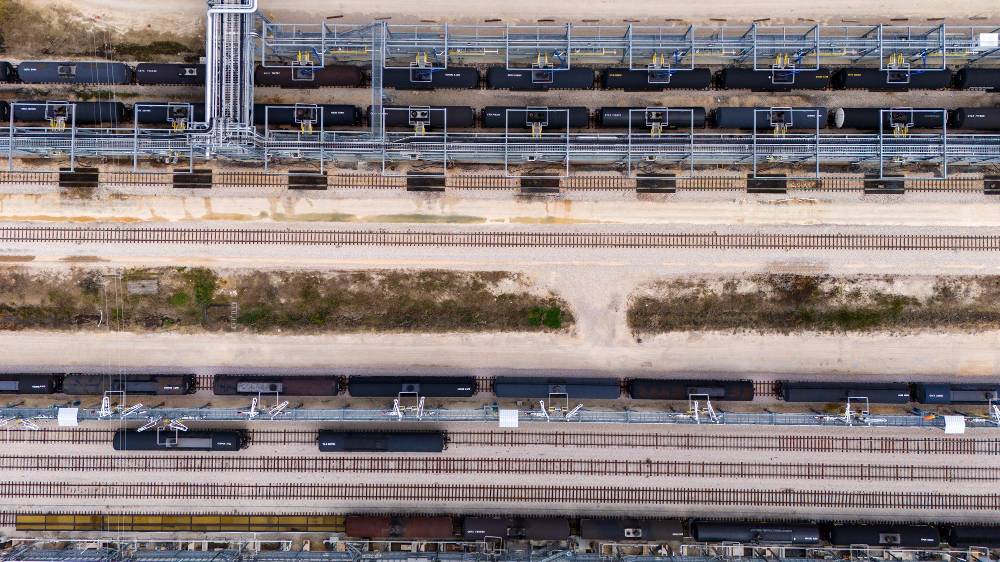 An aerial view of multiple train tracks with several black freight trains and tank cars, some with pipes and industrial equipment, in a rail yard.