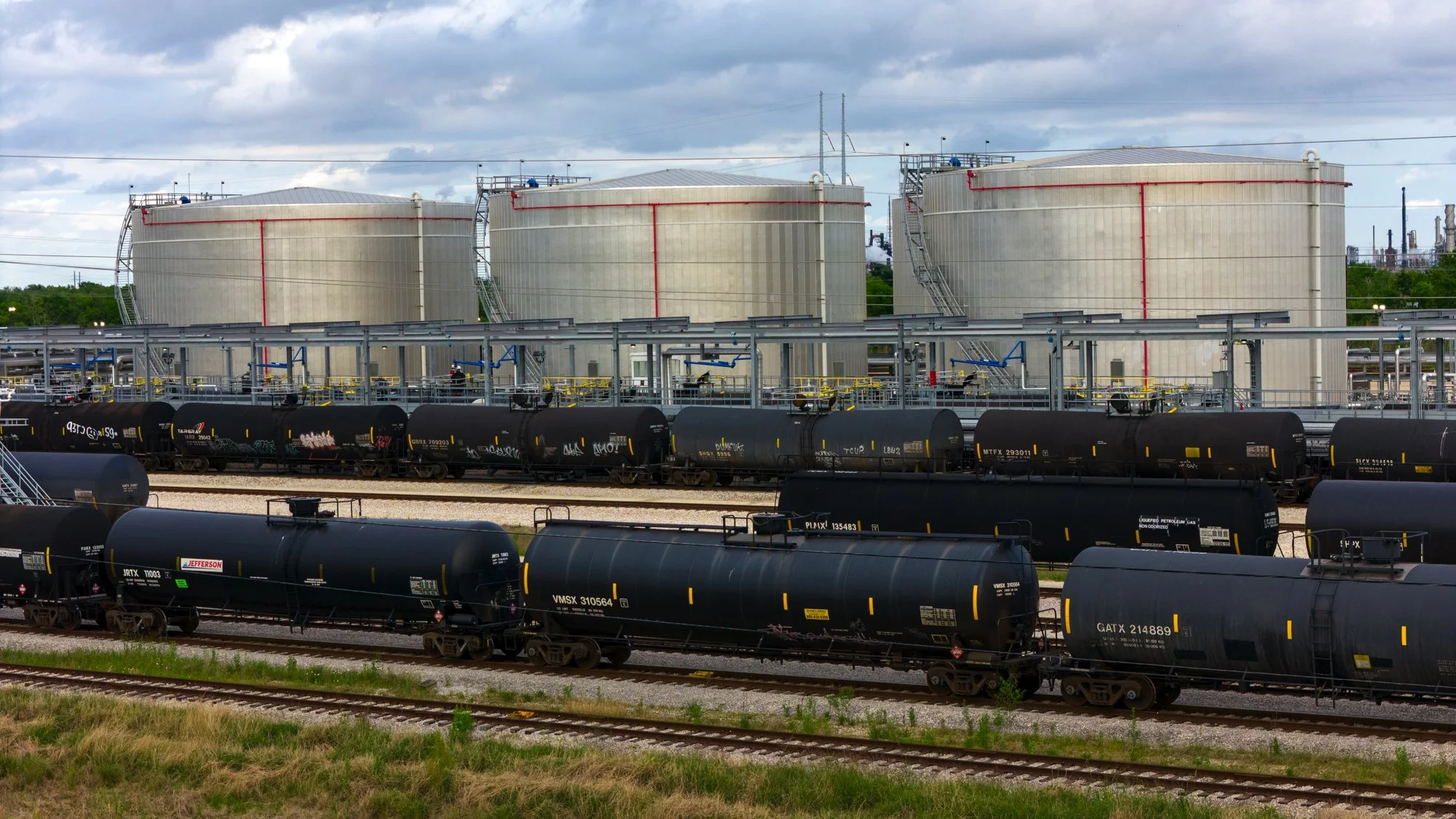 Multiple black tank cars on railway tracks in front of large white storage tanks at an industrial facility.