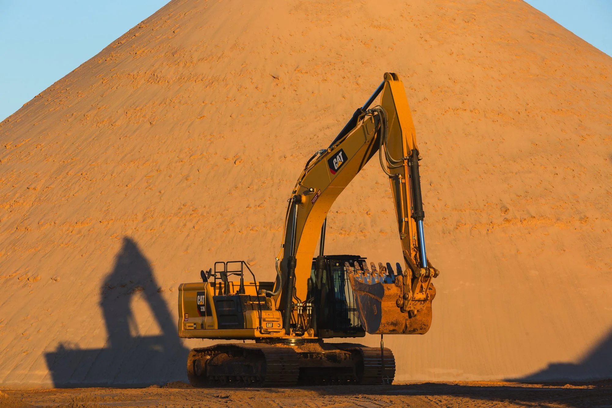 A yellow Caterpillar excavator with a scooping bucket parked in front of a large sand or dirt mound, casting a shadow on the ground during sunset or sunrise.