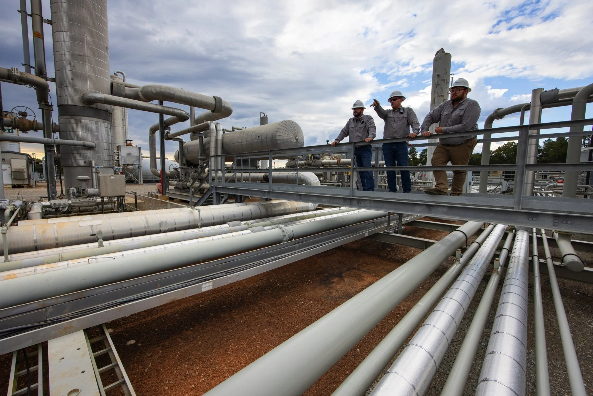 Three industrial workers in grey shirts and hard hats standing on a metal platform inspecting a large industrial facility with many pipes and tanks.
