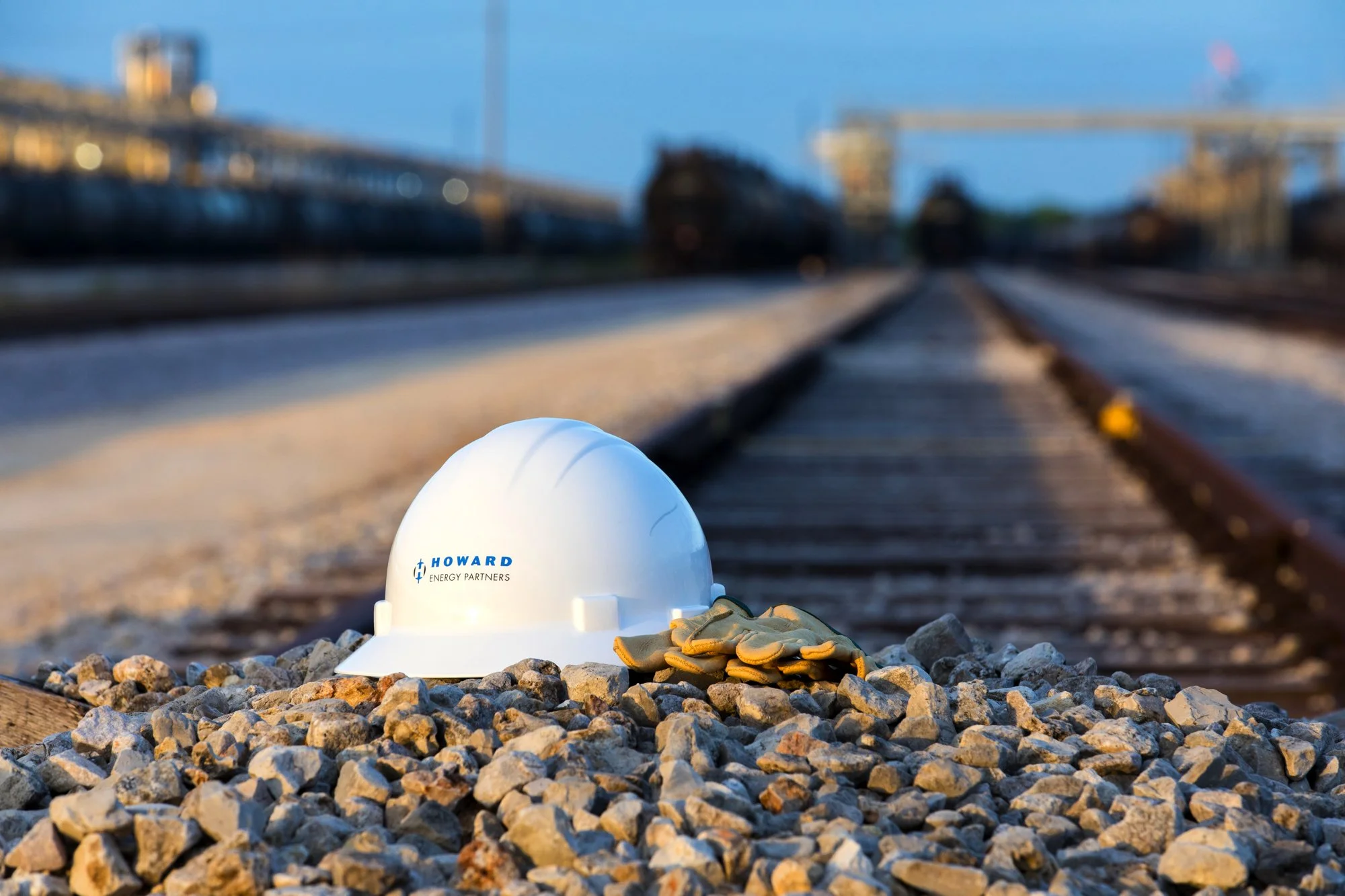 White hard hat with 'Howard Energy Partners' logo and a pair of work gloves resting on rocks beside train tracks in an industrial rail yard.