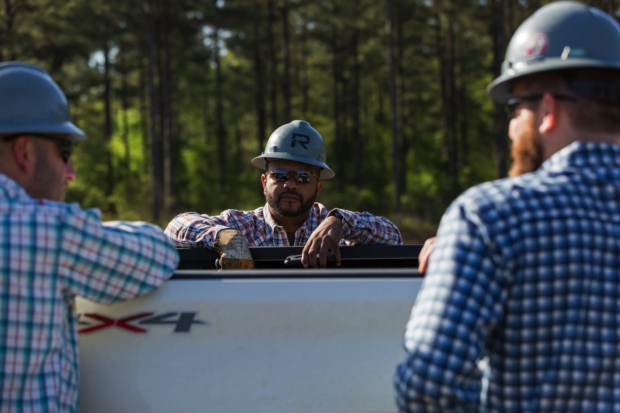 Three workers wearing hard hats and plaid shirts, standing outdoors in a wooded area, having a discussion around a utility or construction vehicle.