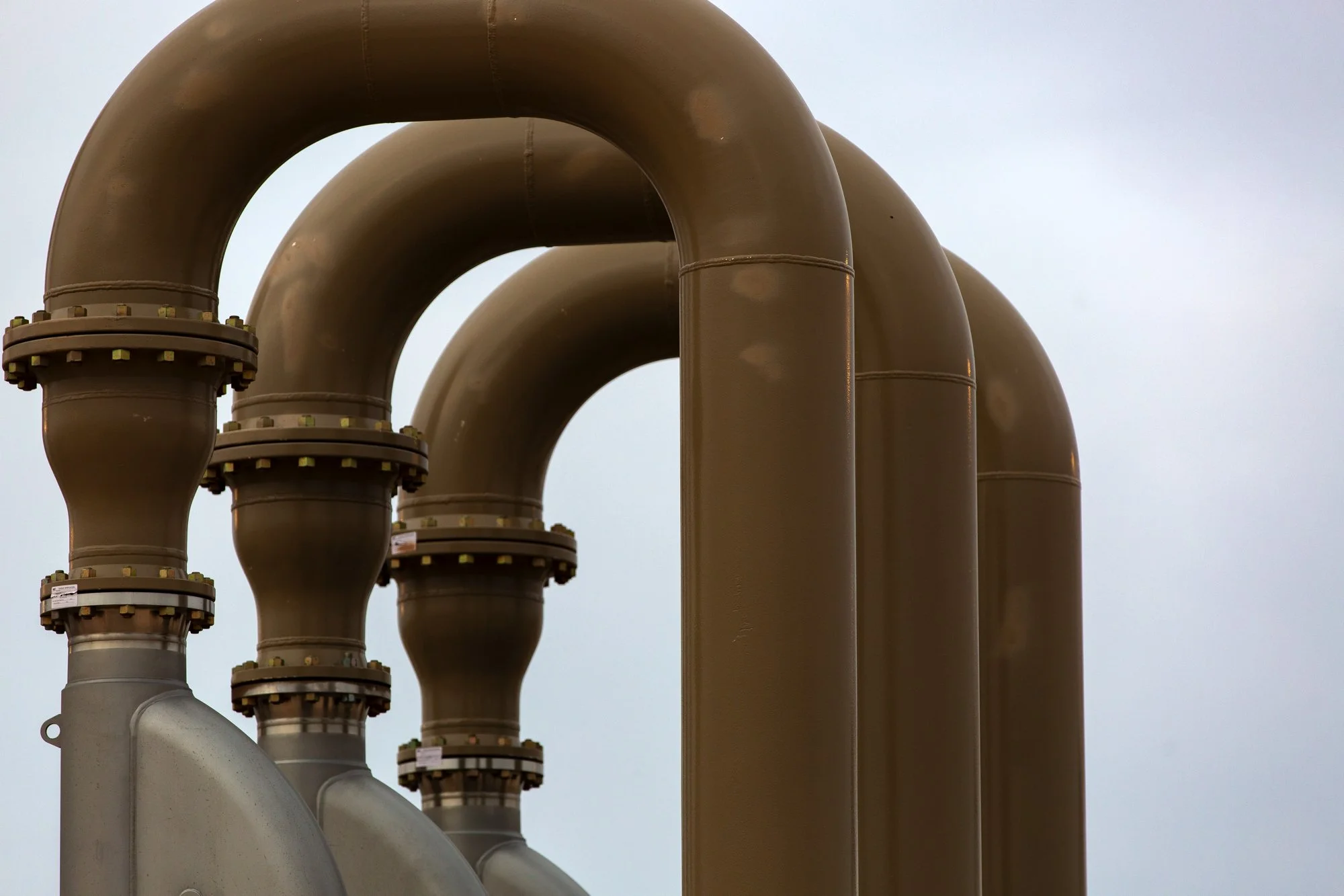 Multiple large brown industrial pipes with flanged joints against a cloudy sky