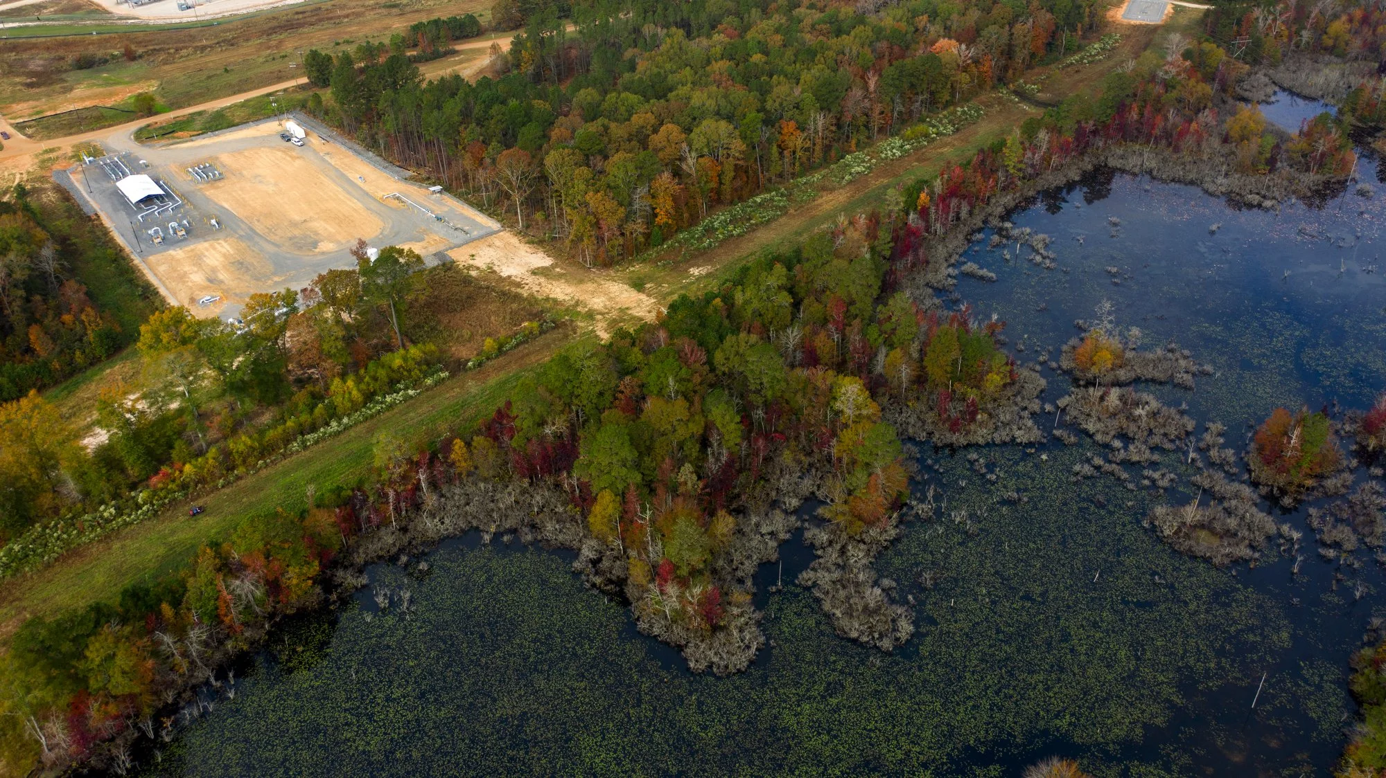 Aerial view of a lake with trees in fall colors, nearby construction site with dirt roads, and a wooded area.