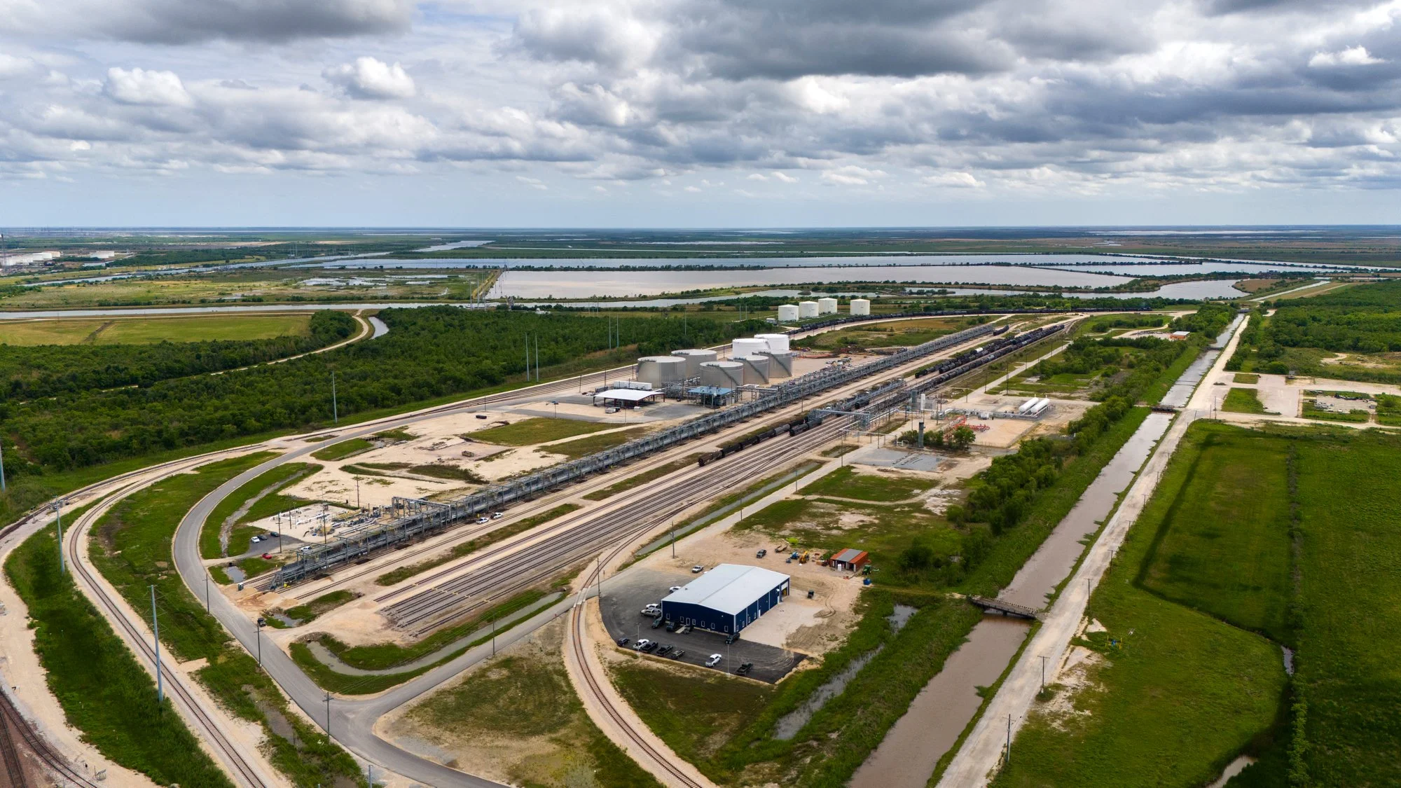 Aerial view of a freight train on multiple tracks passing through green fields and industrial buildings with tanks and warehouses, under a cloudy sky.