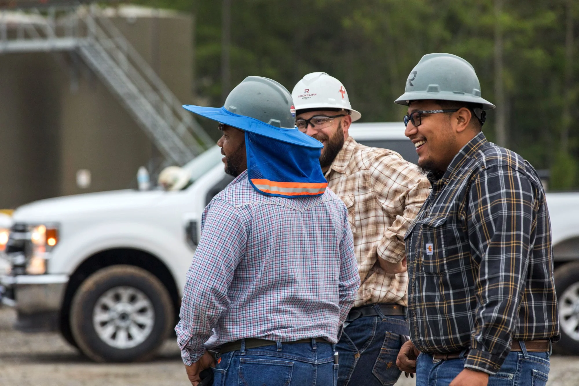 Three construction workers wearing hard hats and plaid shirts, standing outdoors and engaging in conversation, with trucks and construction site elements in the background.