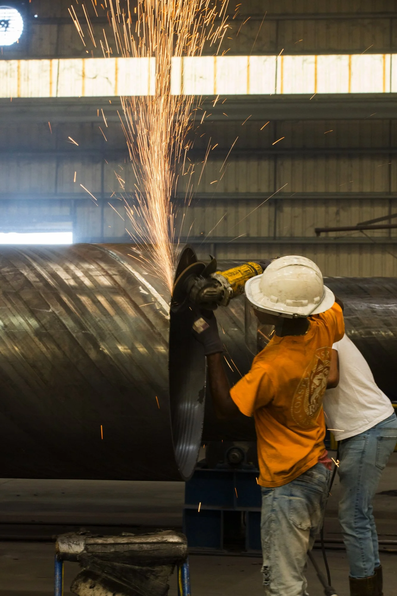 Two workers in safety gear welding a large metal pipe inside an industrial warehouse, with sparks flying from the welding process.