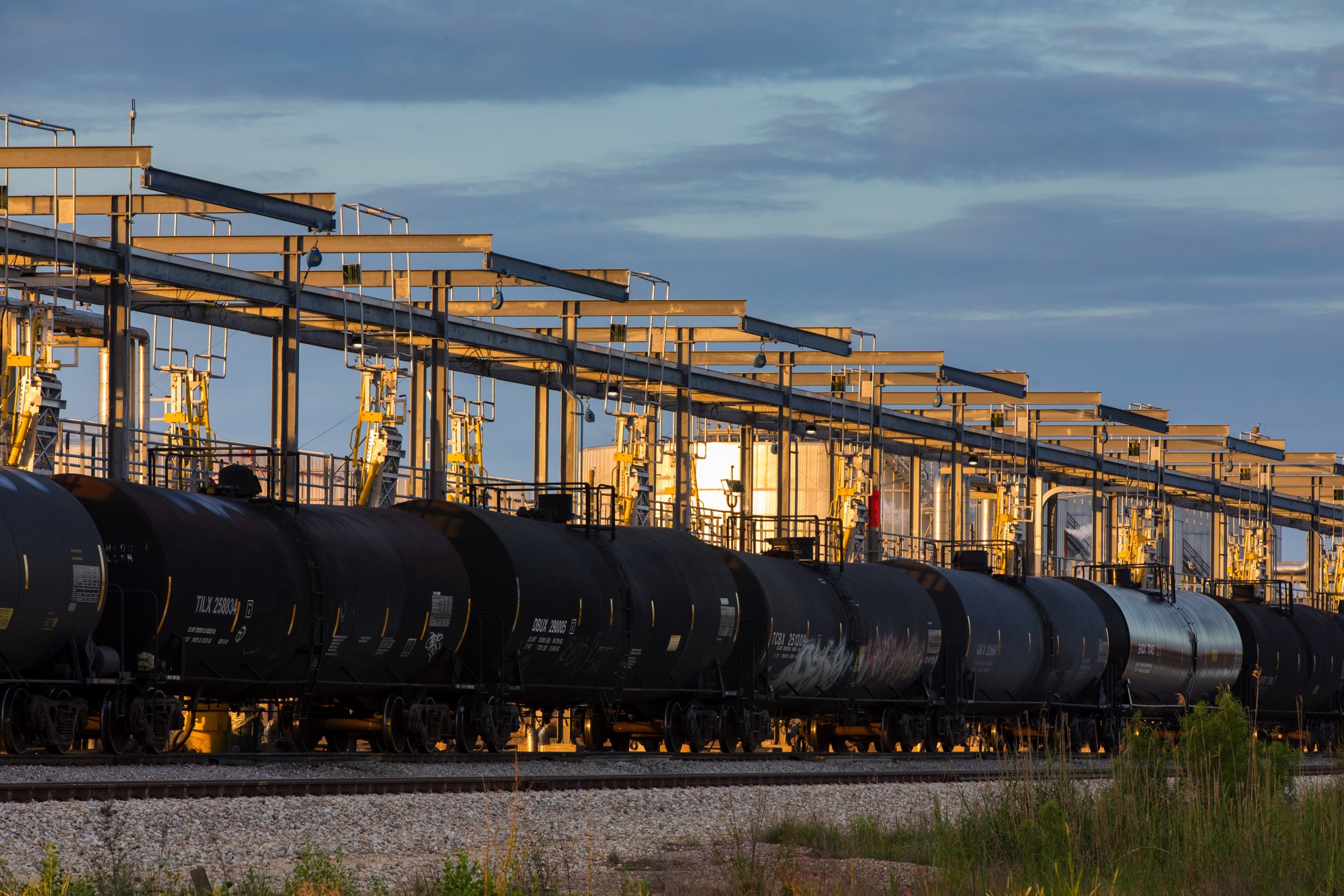 A row of black tanker trains on railway tracks in front of industrial structures with yellow equipment, under a cloudy sky.