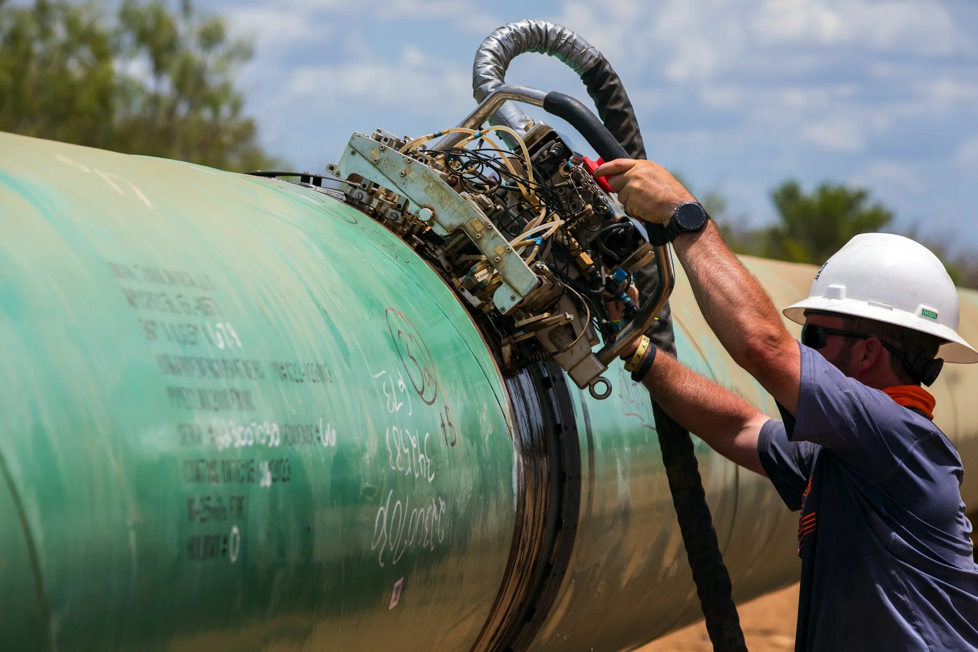 An engineer wearing a white safety helmet and sunglasses is inspecting and working on the wiring of a large green industrial pipeline outdoors, with a cloudy sky and trees in the background.