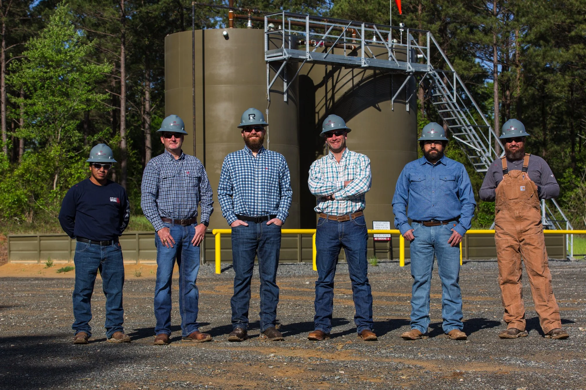 Group of six men standing outdoors in front of industrial tanks, all wearing safety helmets and work boots, with some in casual plaid shirts and jeans.