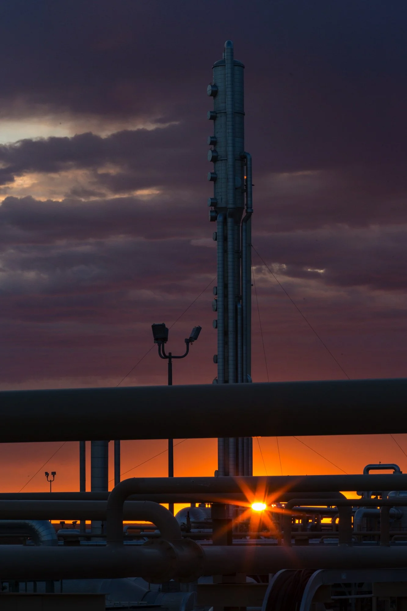 Industrial oil refinery with a tall distillation tower, pipes in the foreground, and a setting sun casting an orange glow behind the equipment, under a cloudy sky.