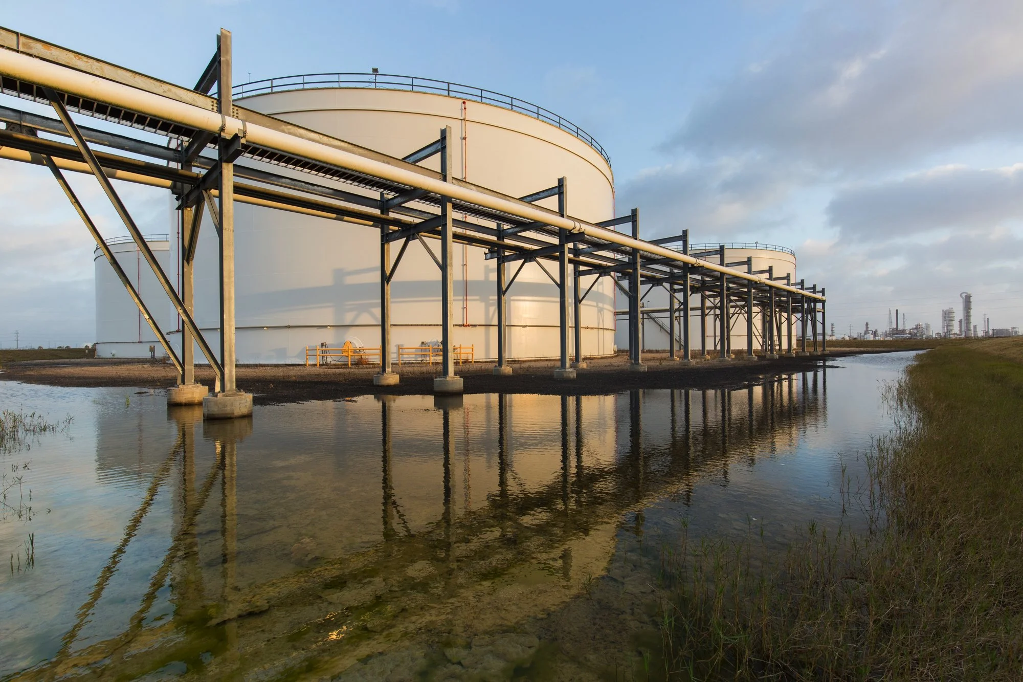 Large white storage tanks at an industrial site with a pipeline structure, reflection in nearby water, and distant industrial facilities under a partly cloudy sky.