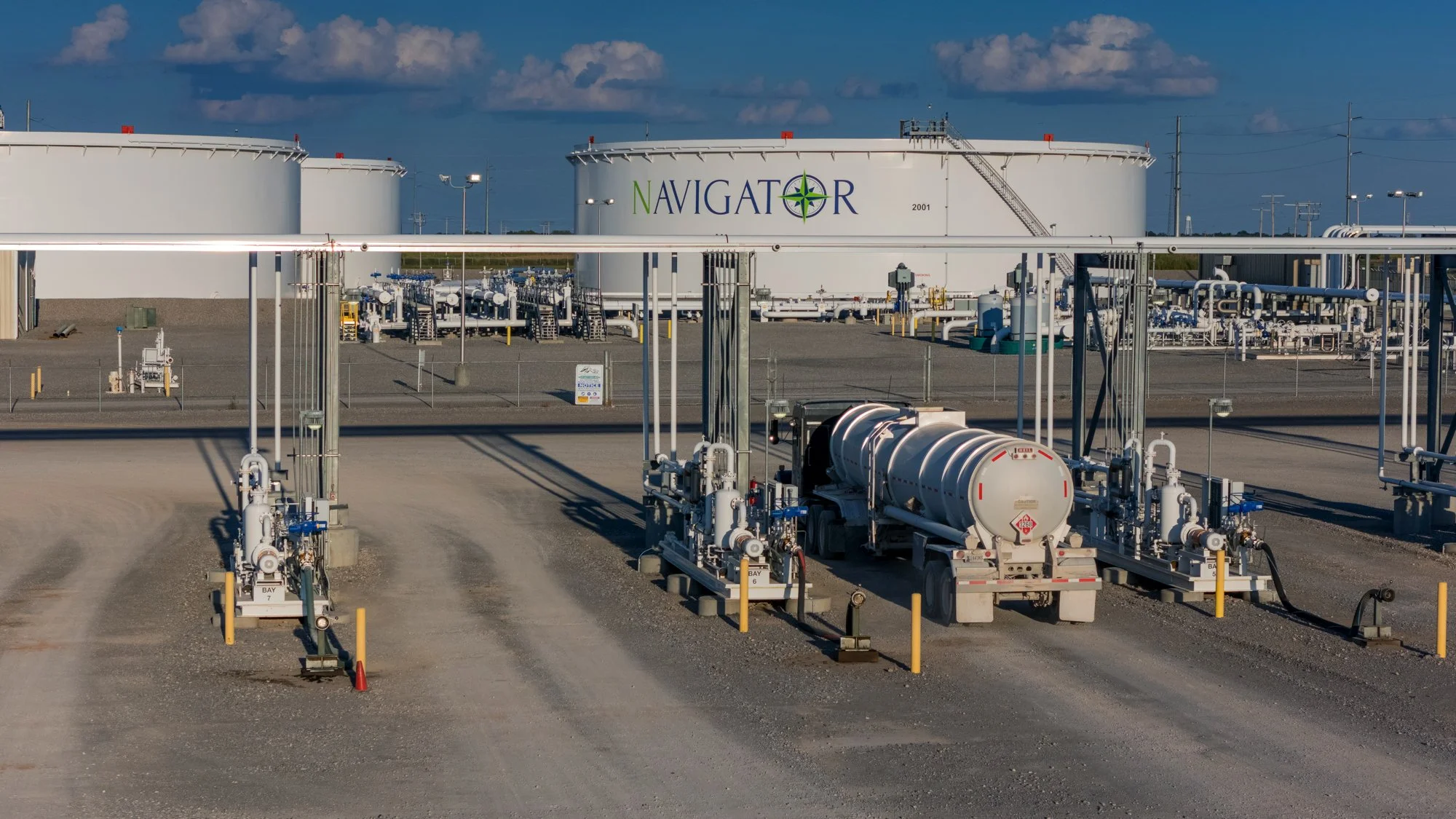 Oil refinery tanks with the word 'Navigator' displayed on one of them, surrounded by pipelines and industrial equipment.